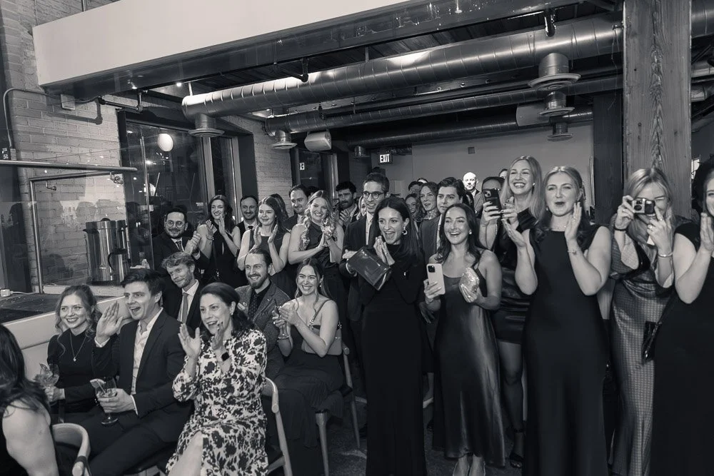 A group of people in formal attire attending an event, some taking photos and videos, smiling and clapping, in an industrial-style venue with exposed pipes and brick walls.