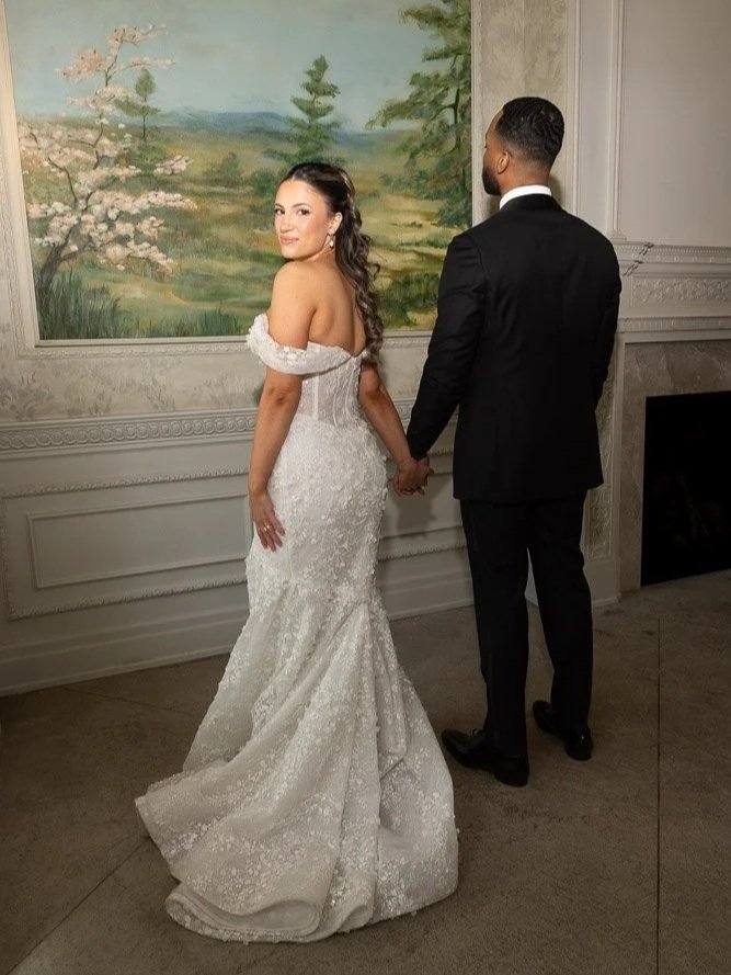 A bride and groom holding hands, standing indoors in front of a landscape painting. The bride wears a white lace wedding gown, and the groom wears a black suit.