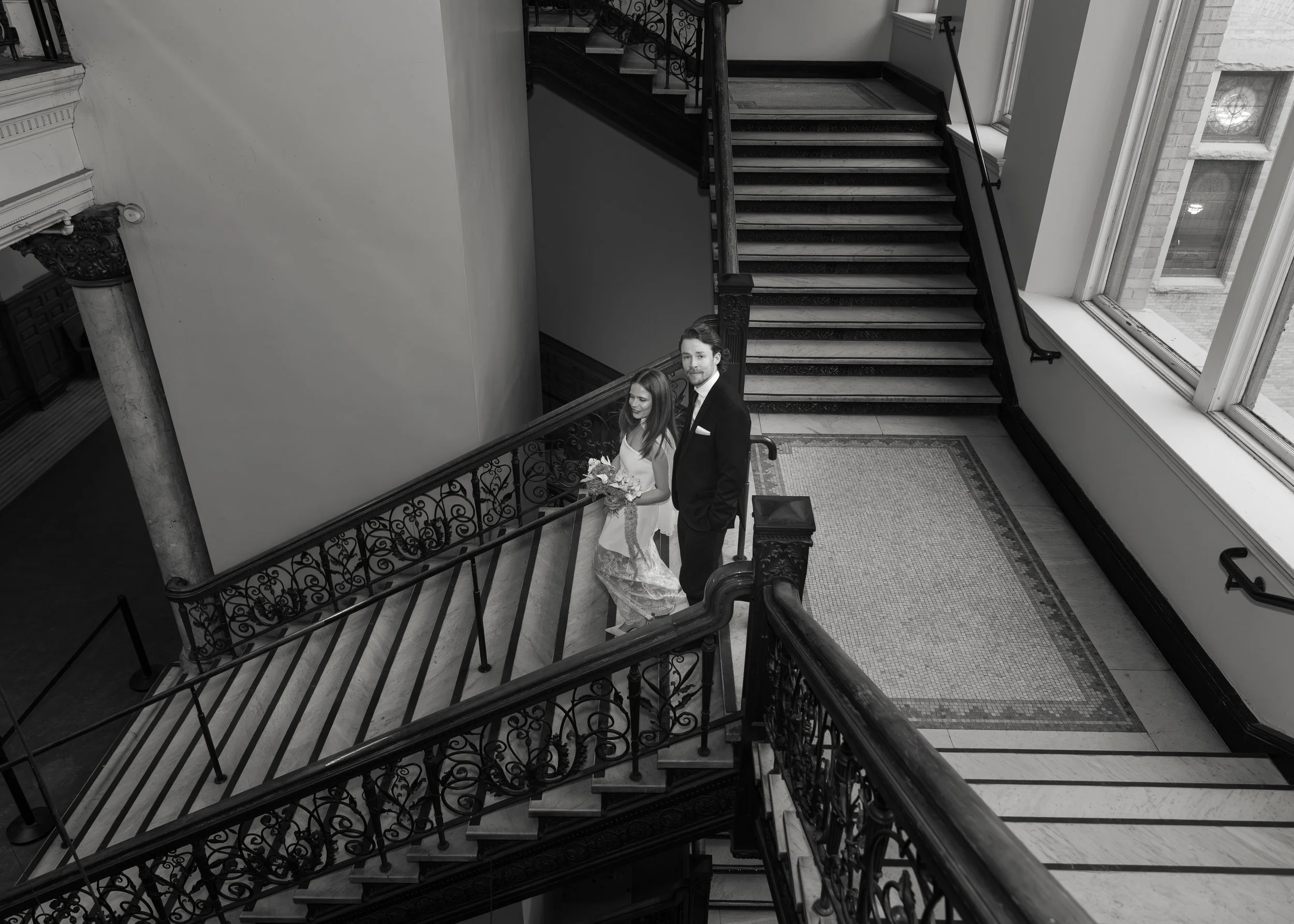 A bride and groom standing on a staircase in a historic building, viewed from above. The bride is holding a bouquet and wearing a wedding dress, while the groom is dressed in a tuxedo, both smiling.