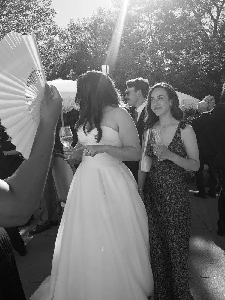 Women at a wedding reception outdoors, one in a white wedding dress, the other in a floral dress, holding wine glasses, with guests and trees in the background.