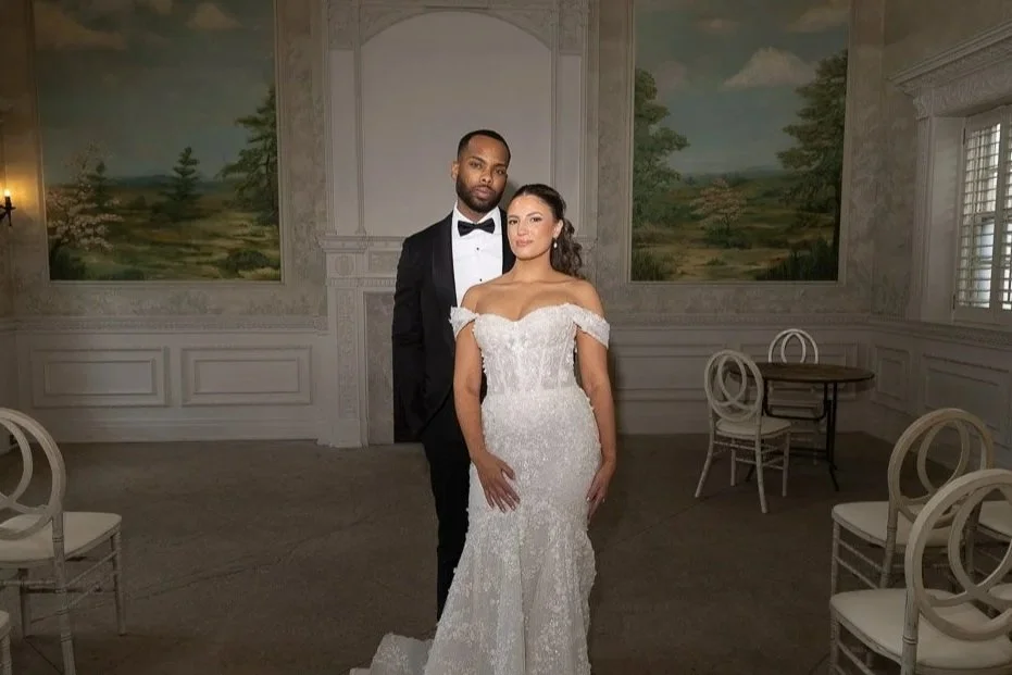 A couple dressed in wedding attire standing in an elegant, decorated room with mural paintings on the walls and white chairs arranged around.