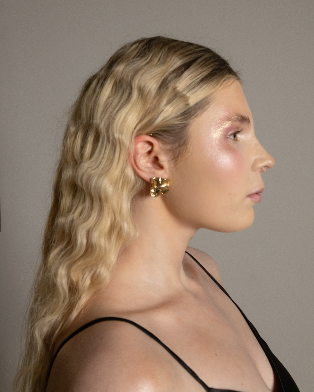 Side profile of a young woman with wavy blonde hair, gold earrings, and natural makeup, wearing a spaghetti strap top against a plain background.