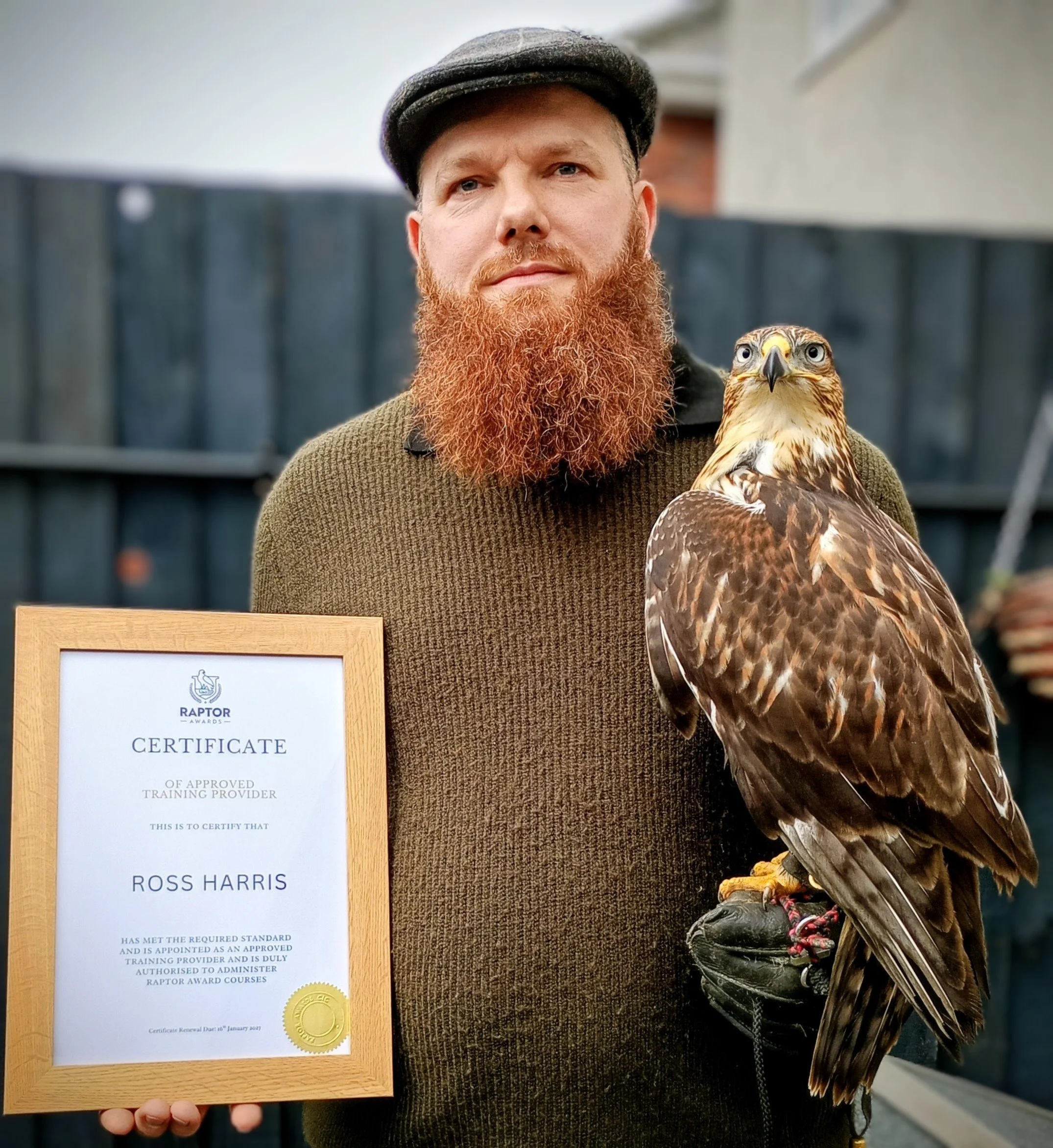 A man with a red beard wearing a black flat cap, holding a brown and white hawk on his arm, standing outdoors in front of a dark wooden fence. The man displays a certificate from Raptor Awards and is standing next to the hawk, which is wearing a leather jess and a anklet.