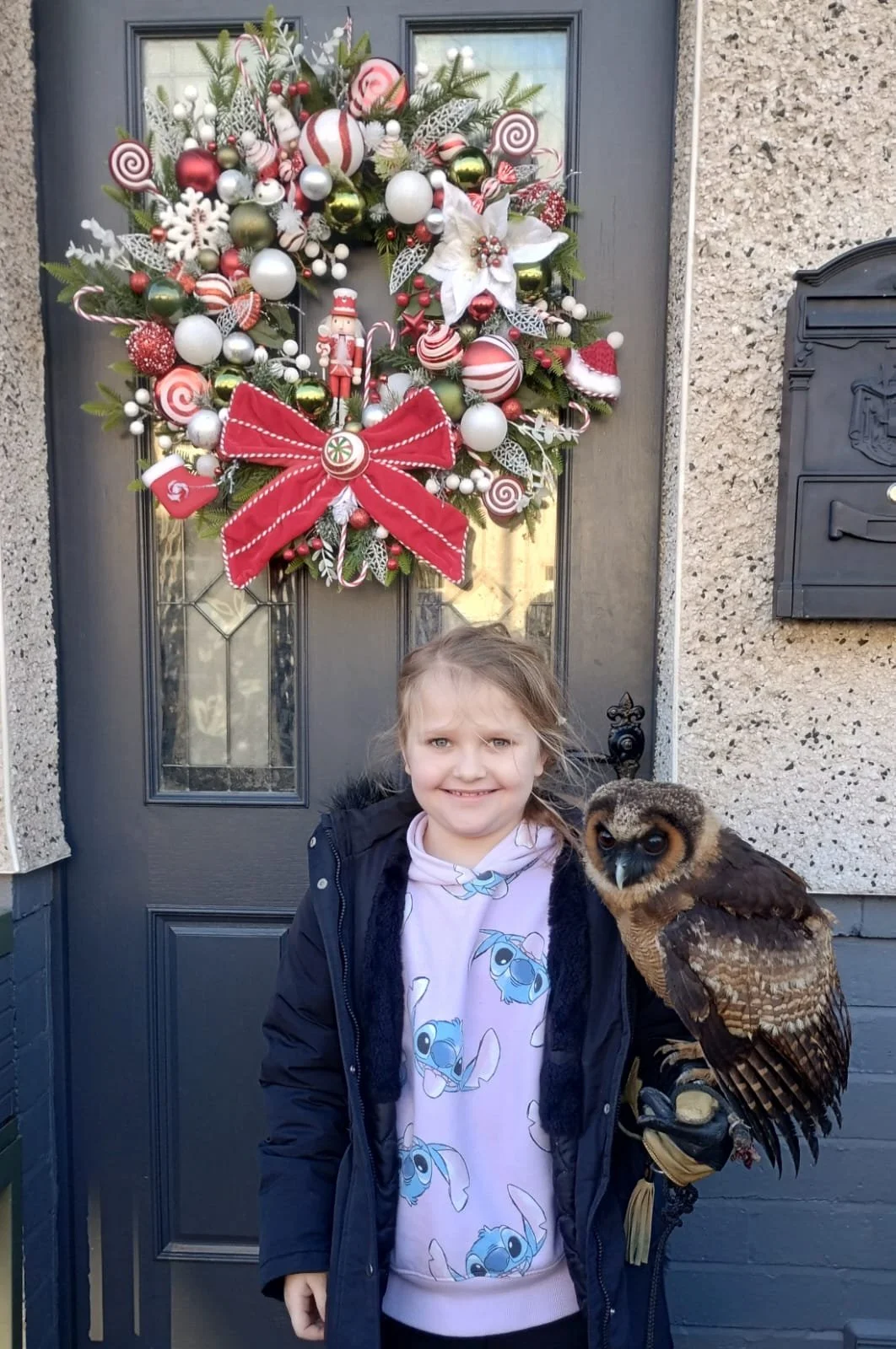 A young girl wearing a Stitch hoodie and a black jacket, holding a large owl, standing in front of a door with a Christmas wreath.