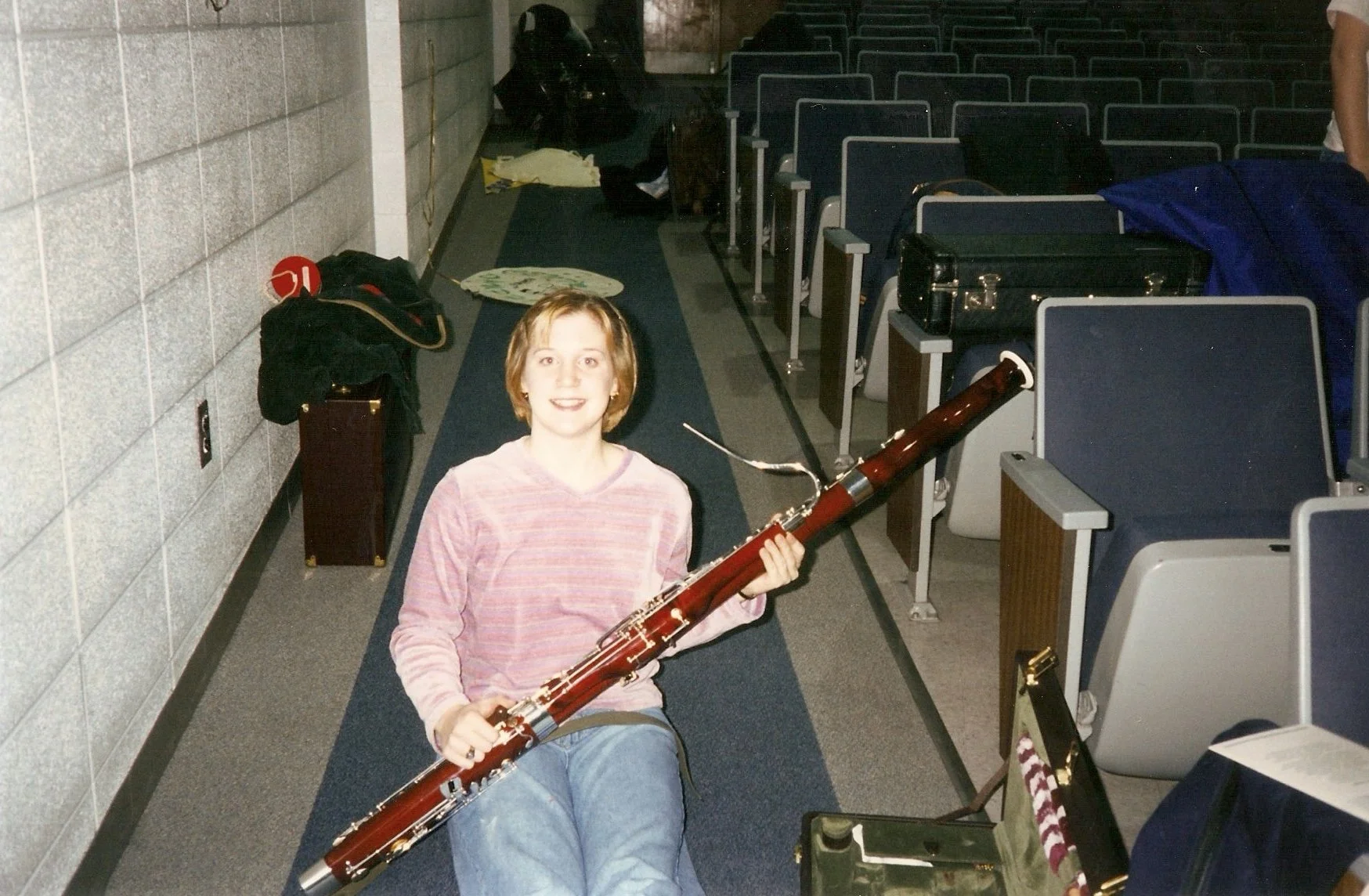 Young girl sitting on the floor at an auditorium, holding a bassoon and smiling at the camera. She is wearing a pink striped shirt, and there are chairs and musical equipment around her.