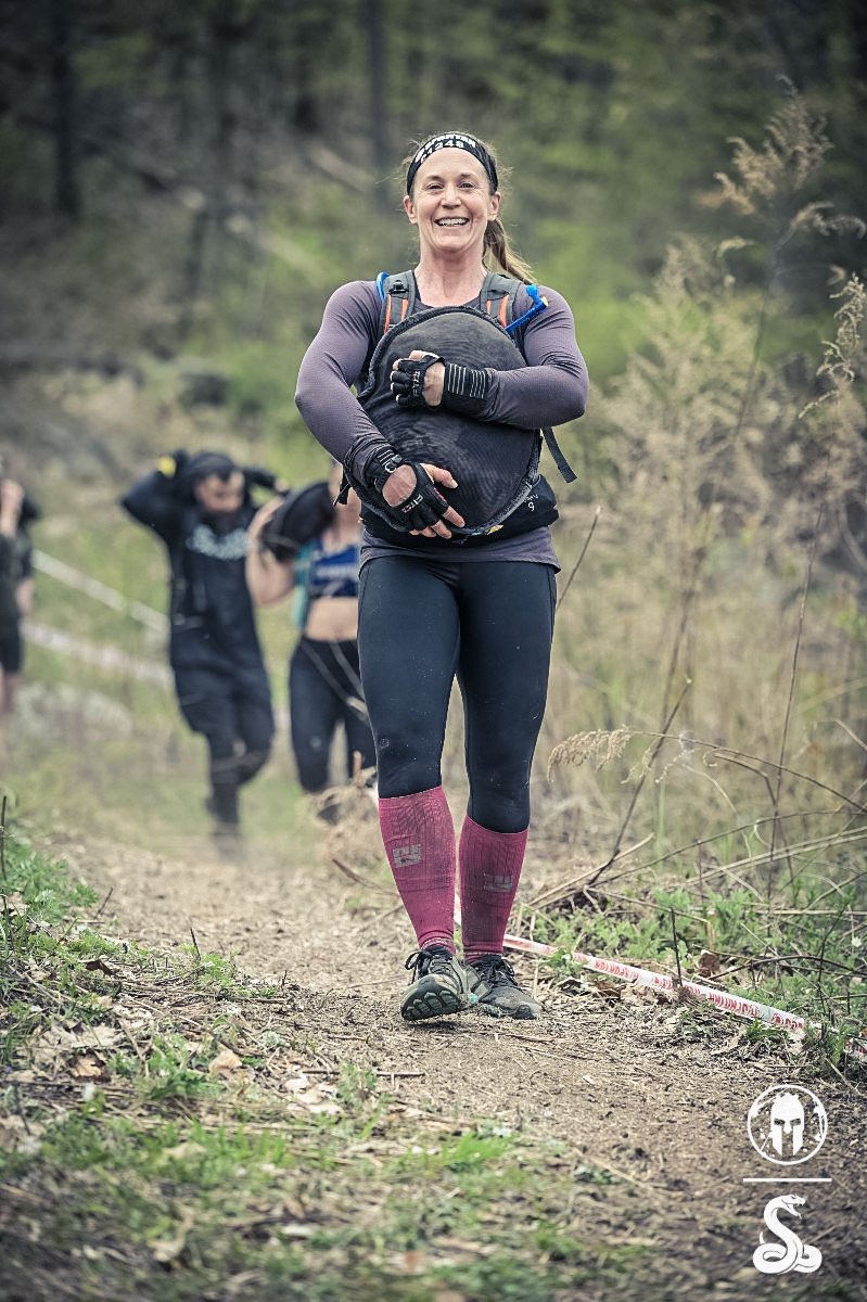 A group of women trail running on a dirt path in a forest, with the woman in the foreground smiling and holding a backpack.