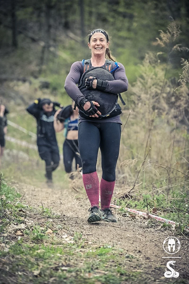 A woman smiling while running on a trail in a forest. She is wearing a headband, long-sleeve athletic shirt, leggings, and pink knee-high socks. Other runners are visible in the background.