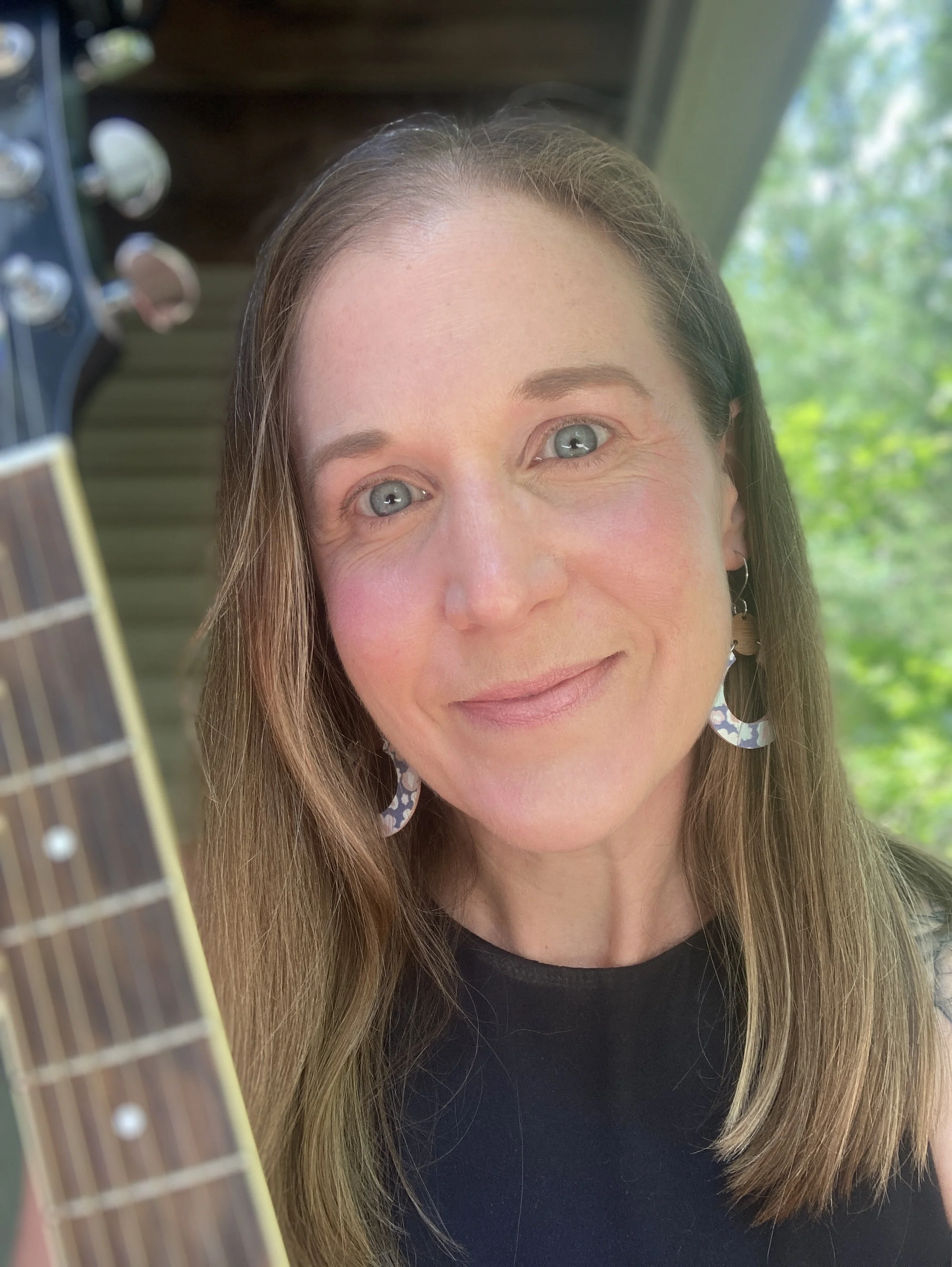 Close-up of a woman with long light brown hair, blue eyes, wearing large patterned earrings and a black top, smiling outdoors with green trees and a window in the background.