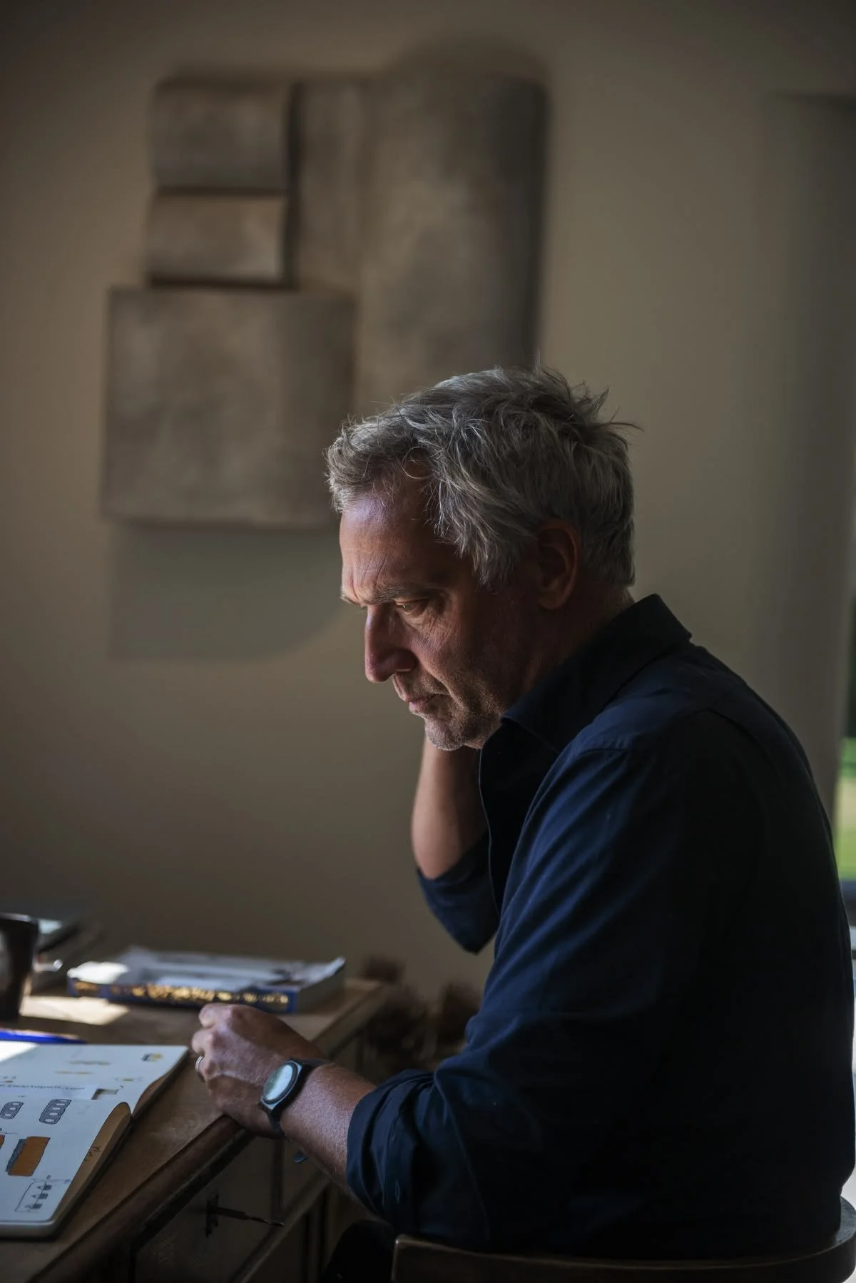 A middle-aged man with gray hair, wearing a dark shirt, is sitting at a desk, looking down and reading or working with papers and a book. The background features a beige wall with an abstract, textured wall art piece.