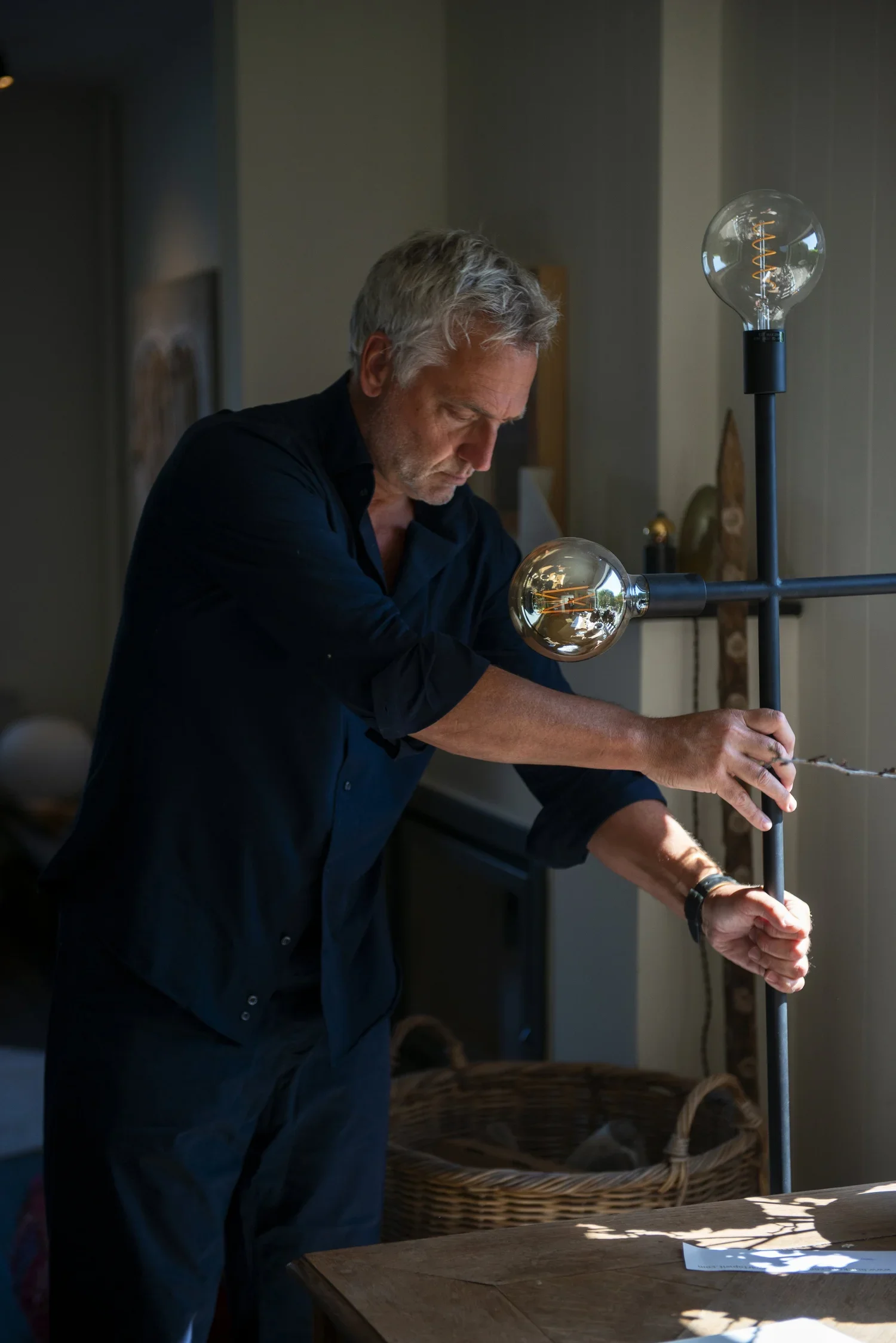 A man with gray hair wearing a black shirt working on an art project at a table. There is a tall lamp with a visible light bulb that has an decorative filament next to him. A basket and paper are also on the table, and sunlight is casting shadows.