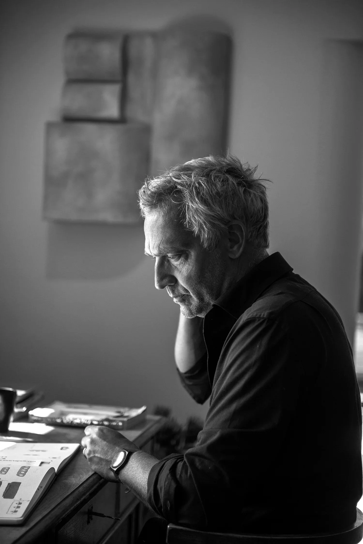 A man with gray hair, wearing a watch, sitting at a desk with books and papers, looking down at his phone or device in a study or work environment.