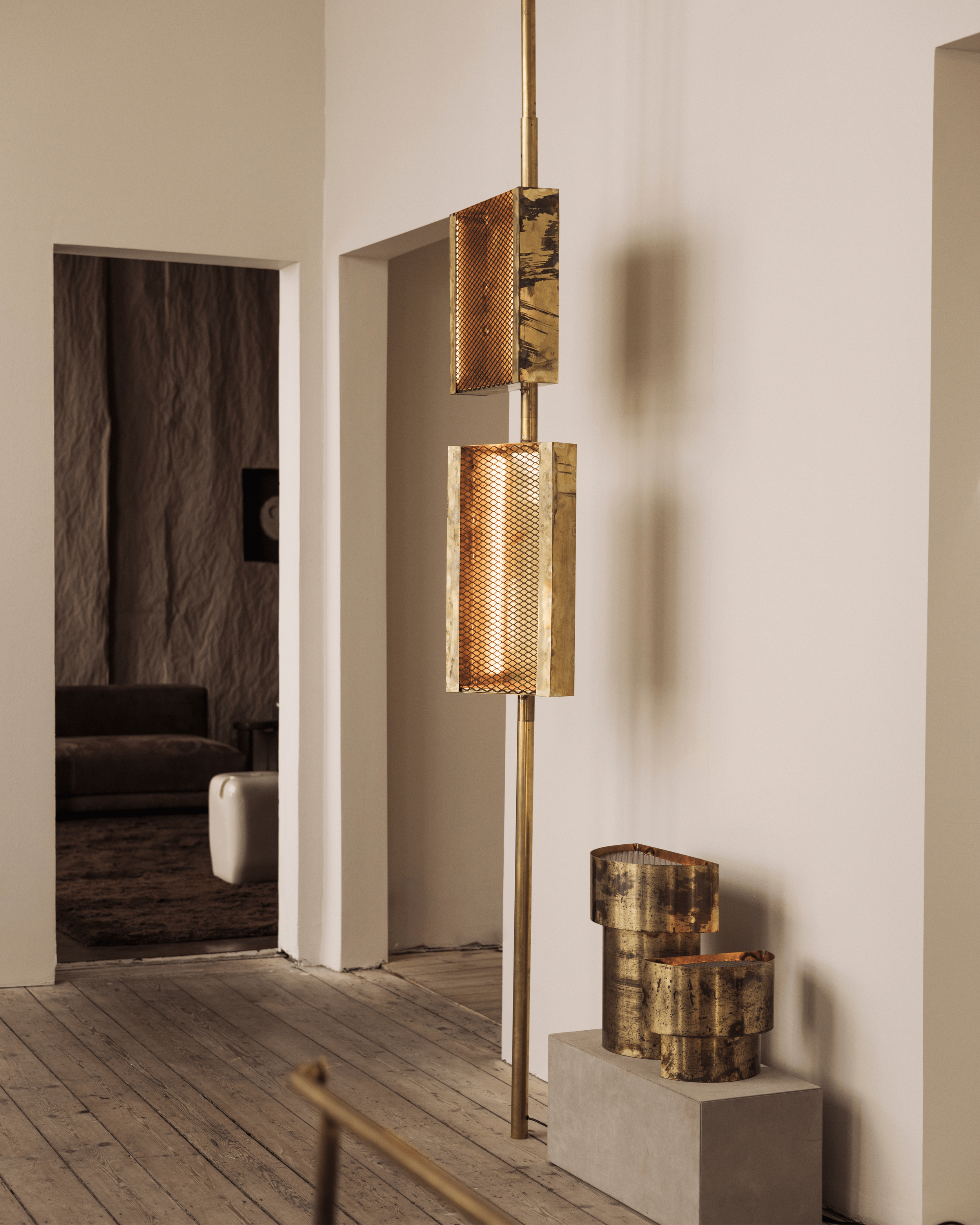 A modern art sculpture made of three cylindrical metal pieces with a gold finish, placed vertically on a rectangular concrete pedestal. The sculpture has two rectangular metal panels with a rusty finish, attached to a vertical pole that extends above and below the panels. The sculpture is positioned in a gallery room with wooden flooring and beige walls.