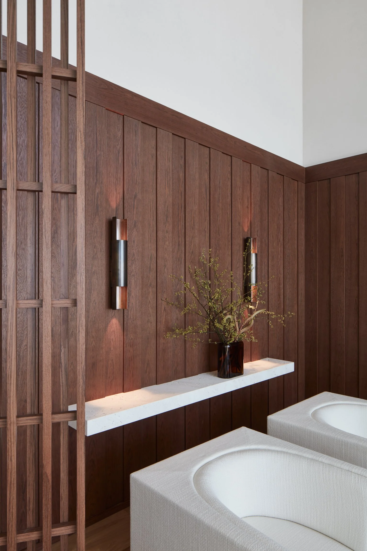 Interior of a room with wood-paneled wall, two wall-mounted lights, a white shelf with a tall glass vase of green foliage, and two white beds in the foreground.
