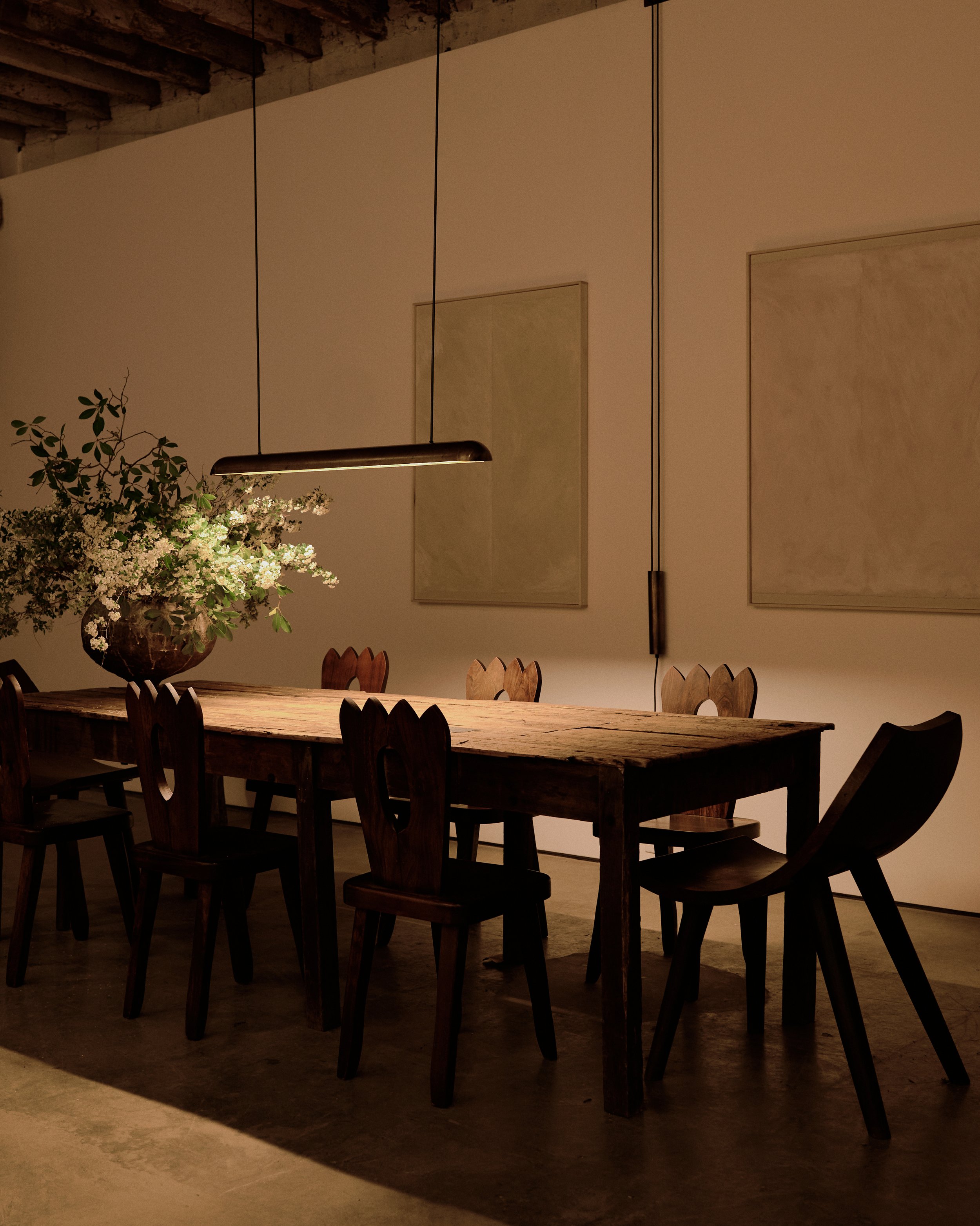 Interior of a modern dining room with a long wooden table and multiple chairs. A large plant with white flowers on the table and a minimalistic pendant light hanging above. The wall features two rectangular pieces of art or mirrors.