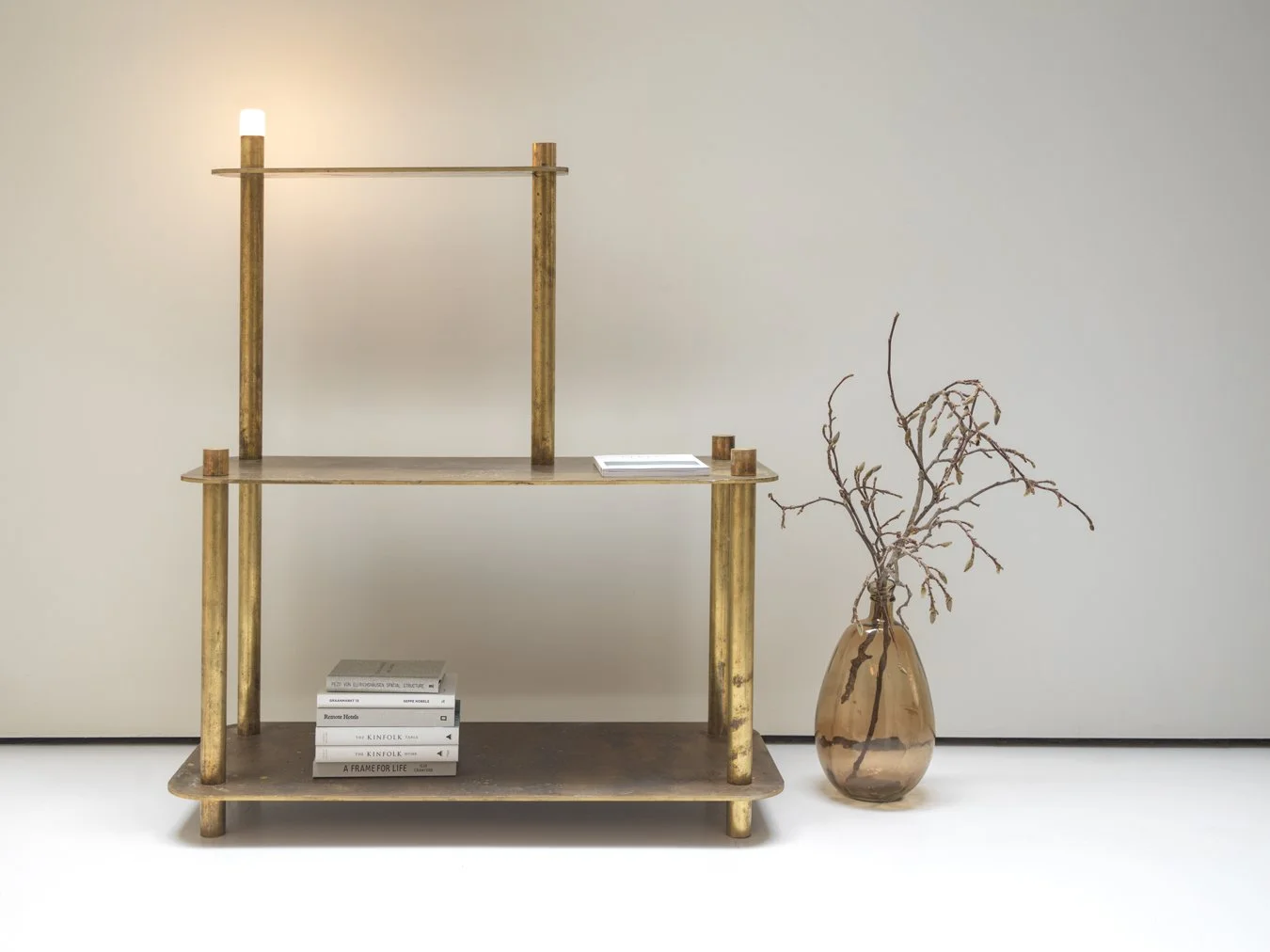 Empty gold-colored display shelf with three levels next to a tall glass vase containing dried, leafless branches, on a plain white background.