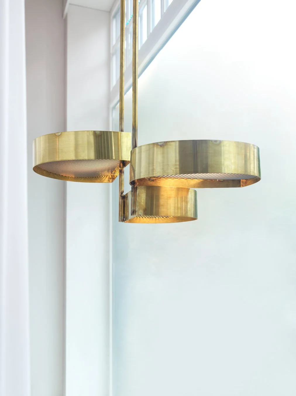 Close-up of a modern brass chandelier with three circular rings, hanging near a window with white framing.