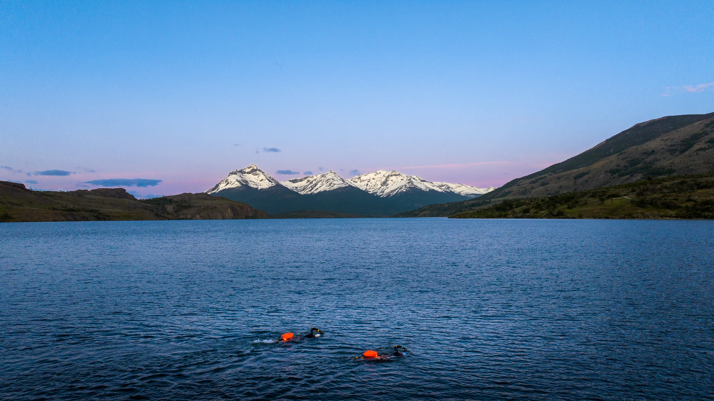 Lago rodeado de montañas con picos nevados bajo cielo despejado y una persona nadando con gorro y aletas en el agua.