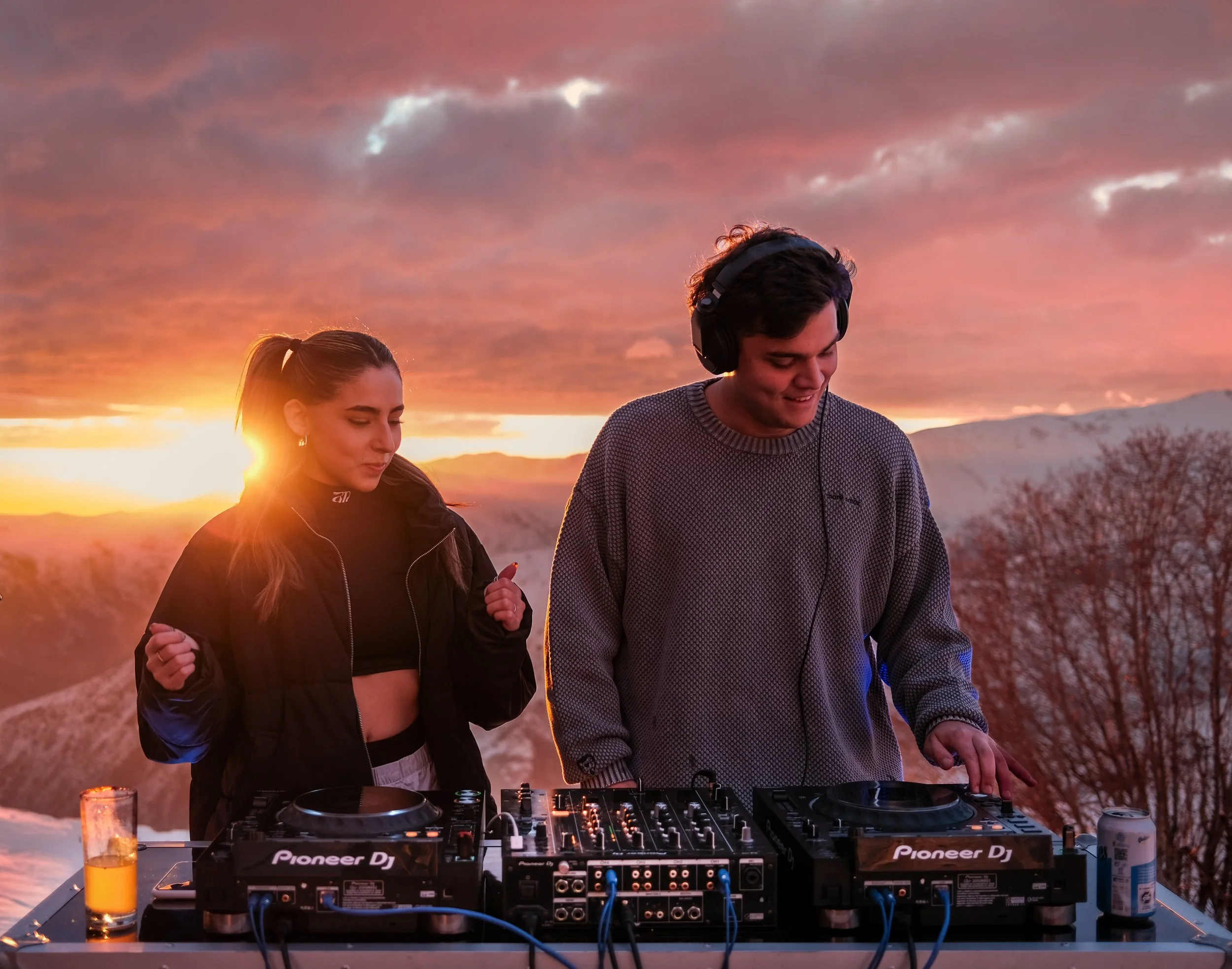 Jóvenes compartiendo música en un paisaje al atardecer, con equipo de DJ en una mesa.
