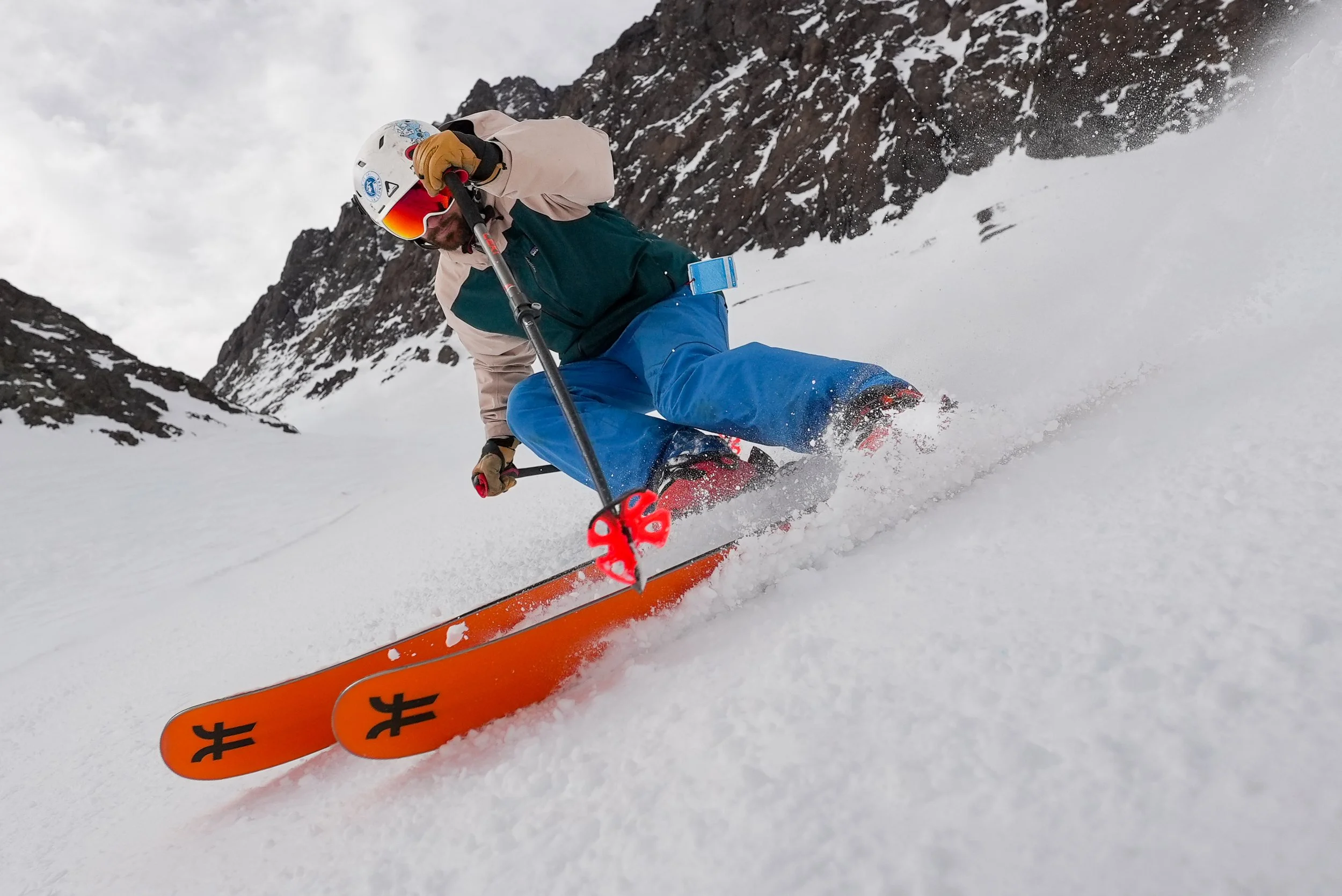 Persona haciendo esquí en una pendiente de nieve en las montañas, usando casco, gafas con visor rojo, y ropa de esquí en tonos beige, verde y azul, con esquís anaranjados.