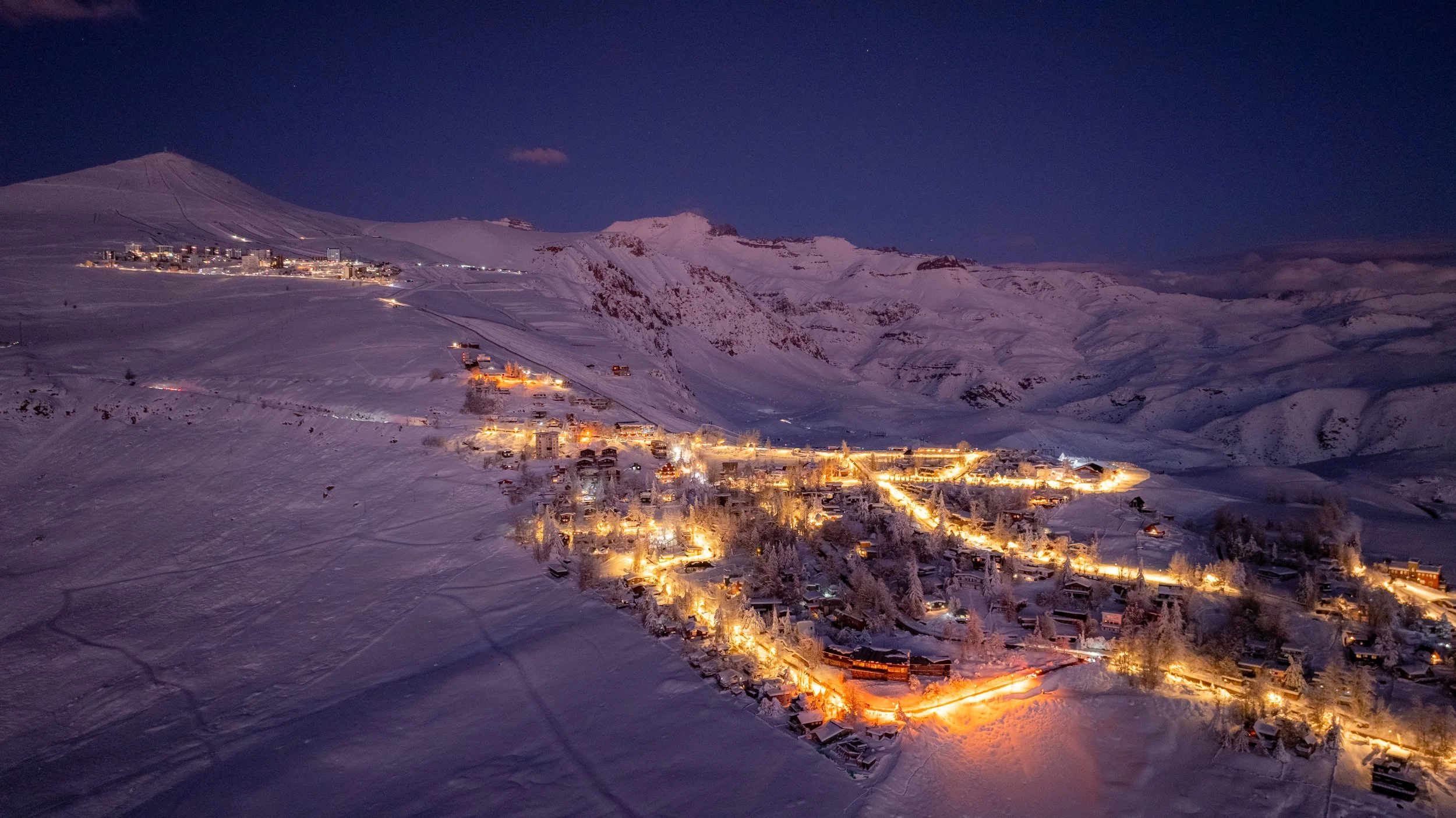 Paisaje nocturno de una ciudad en una montaña cubierta de nieve, con casas iluminadas y un cielo oscuro.