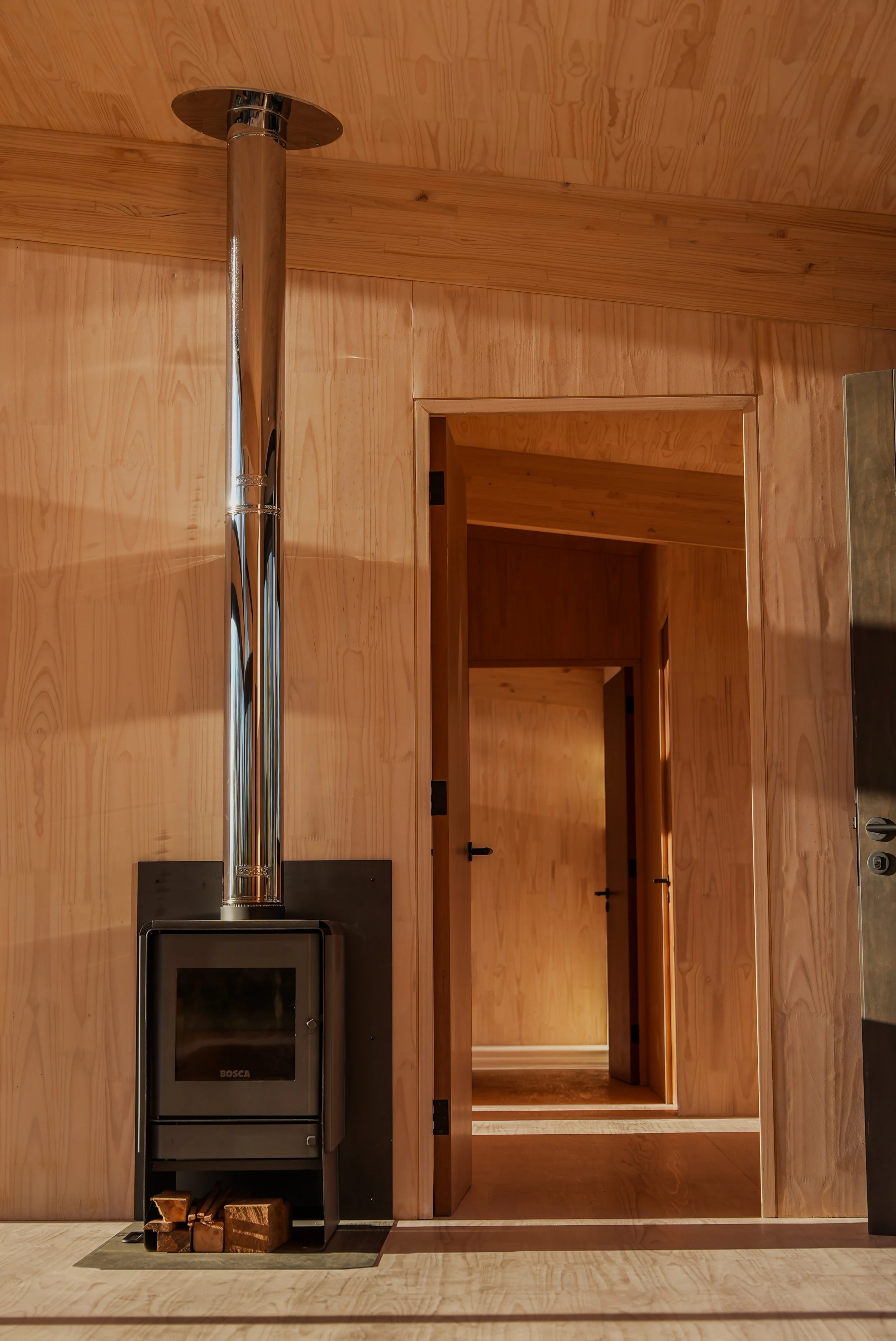 Habitación con paredes y techo de madera, estufa de leña negra con tubo de escape de metal brillante y pila de madera en la base, puerta que lleva a otro espacio con paredes de madera, suelo de madera clara.