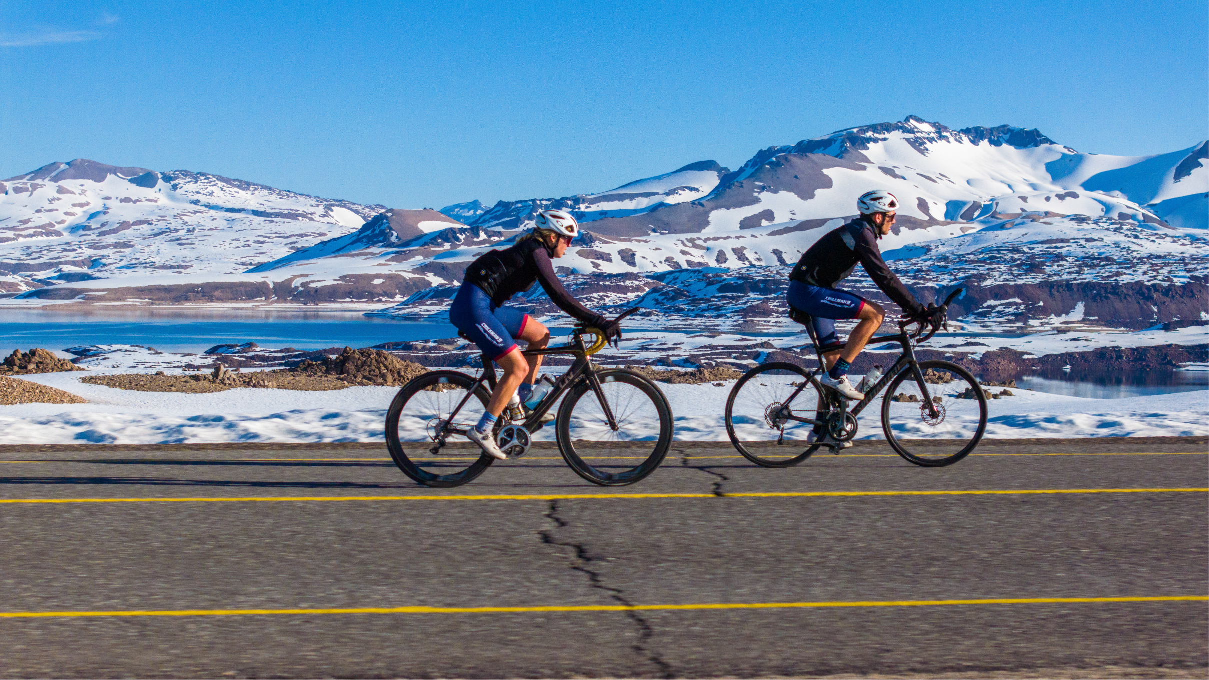 Dos personas en bicicletas pedaleando en un paisaje de montañas con nieve, agua y cielo azul en un día soleado.