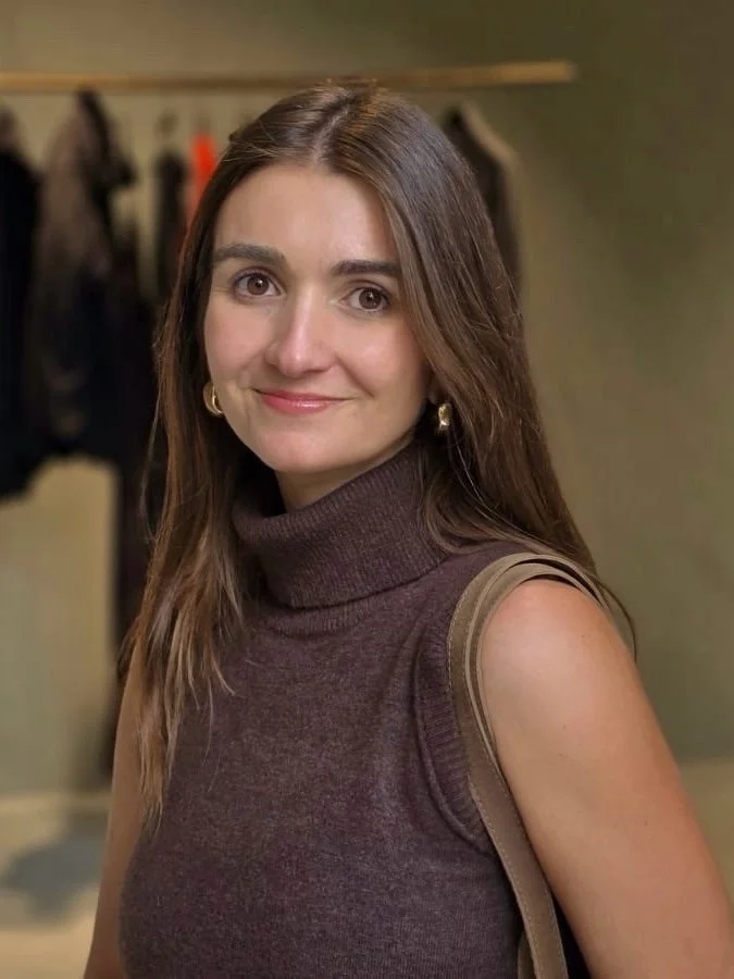 Mujer joven con cabello castaño y ojos marrones, vestida con suéter morado y bolso beige, en interior con fondo borroso
