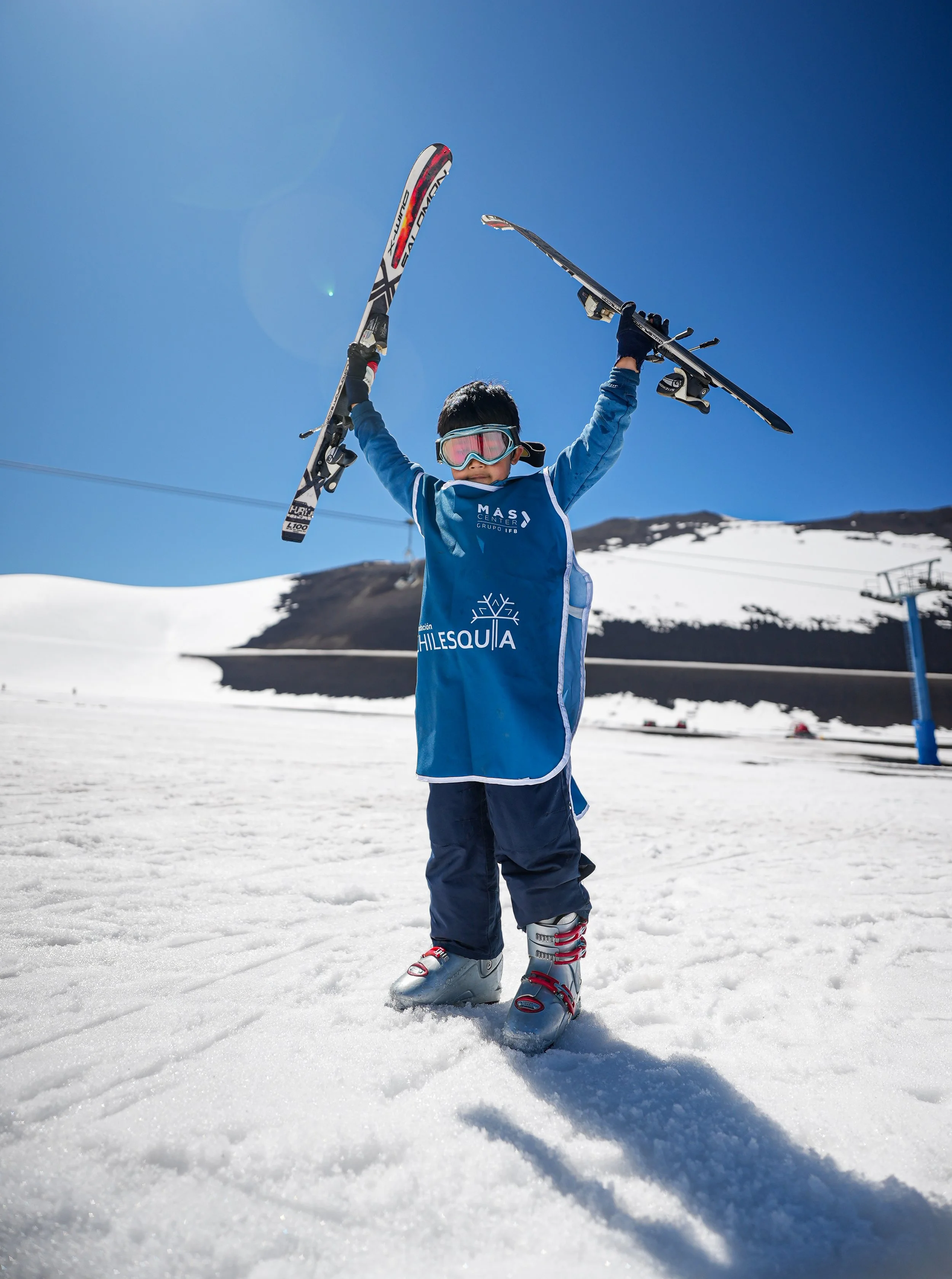 Niño en esquí, vistiendo ropa de invierno y con gafas, levantando los esquís en un paisaje de nieve bajo un cielo azul brillante en una estación de esquí.