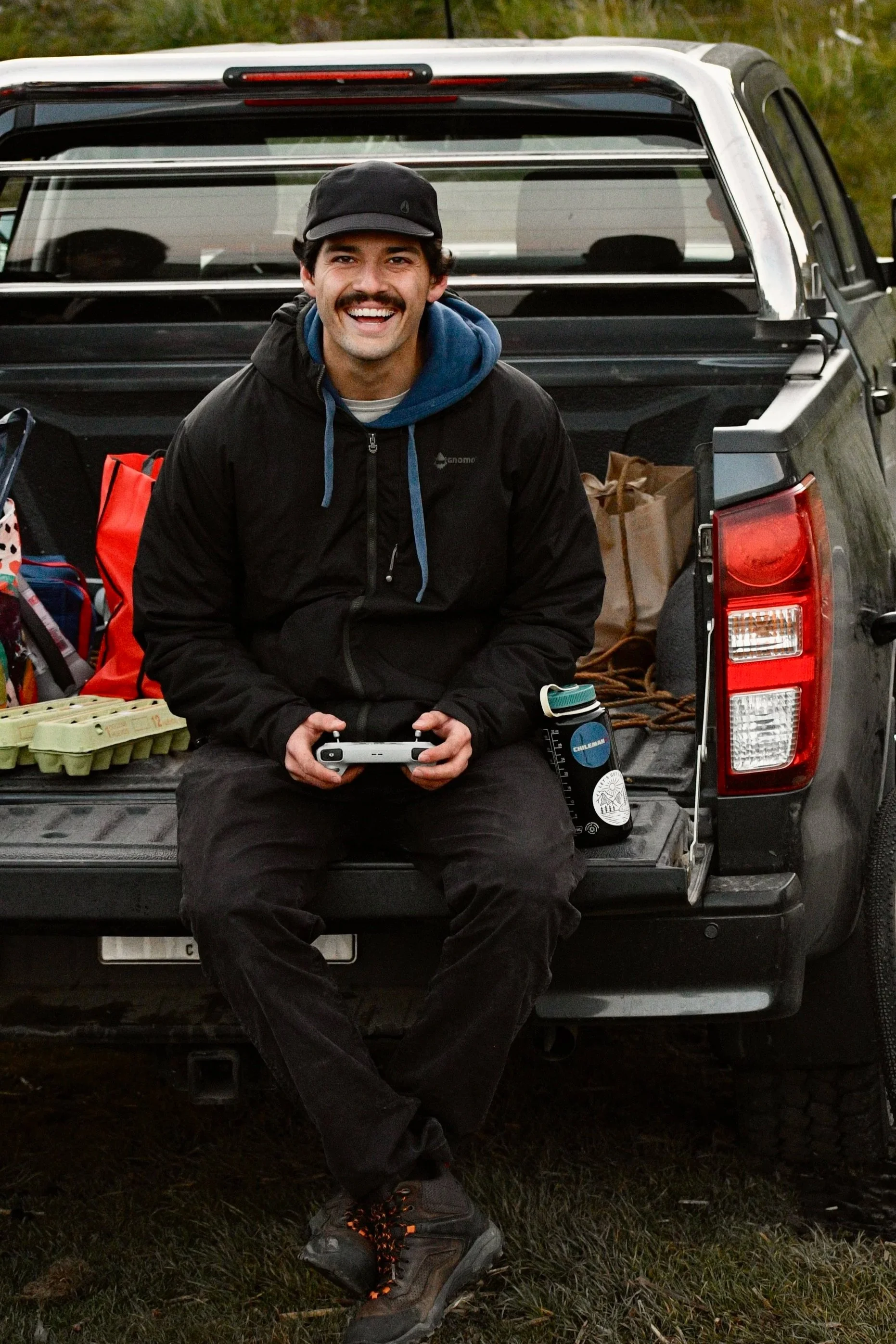 Un hombre sonriente sentado en la parte trasera de una camioneta con equipo de camping, en un entorno de campo o naturaleza.