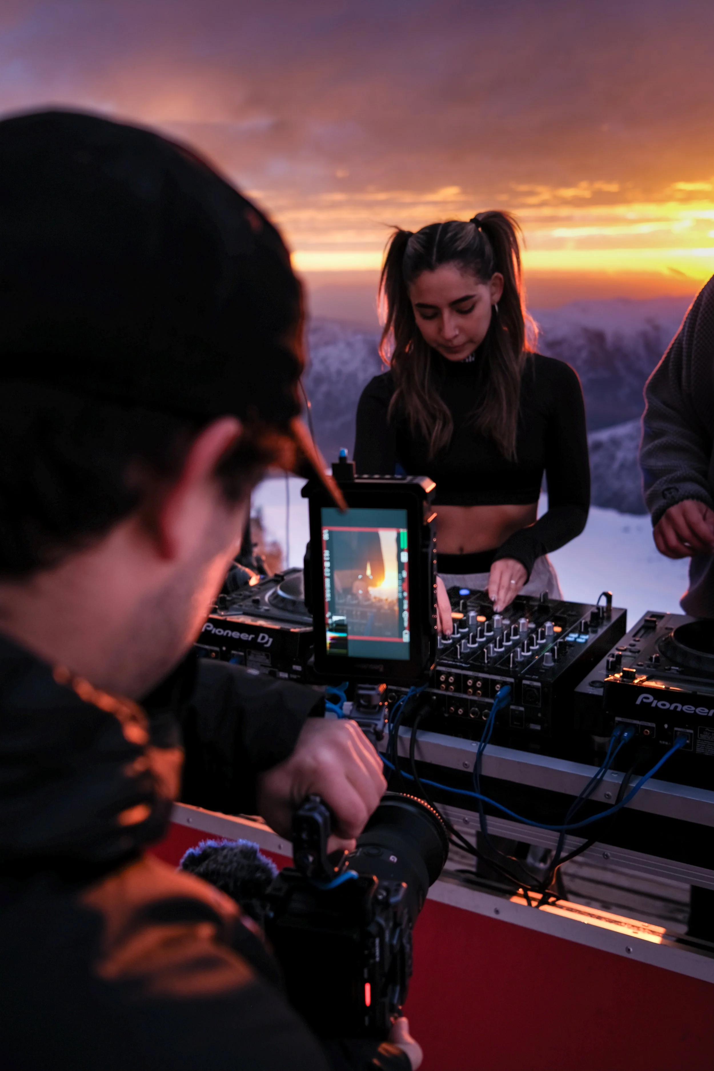 Muchacho con auriculares observa una consola de DJ siendo operada por una mujer en un atardecer con cielo naranja y nubes.