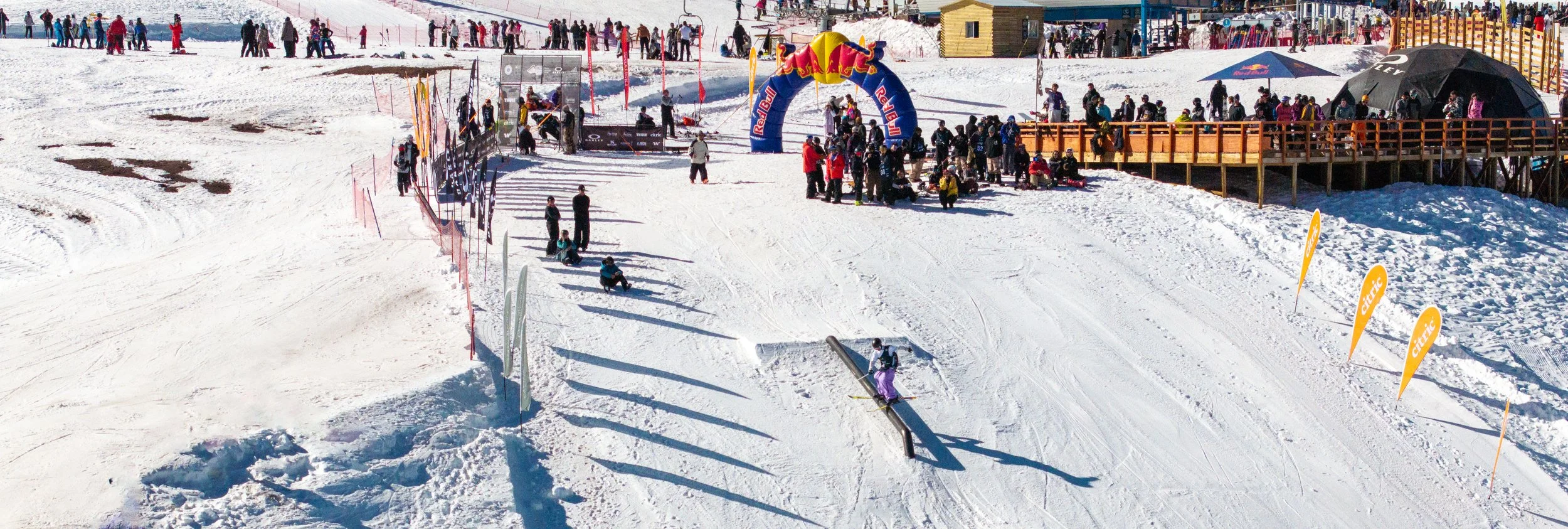 Personas en una pista de esquí en la nieve, con espectadores y estructuras de apoyo, en un día soleado, con banderas y obstáculos para actividades de esquí.