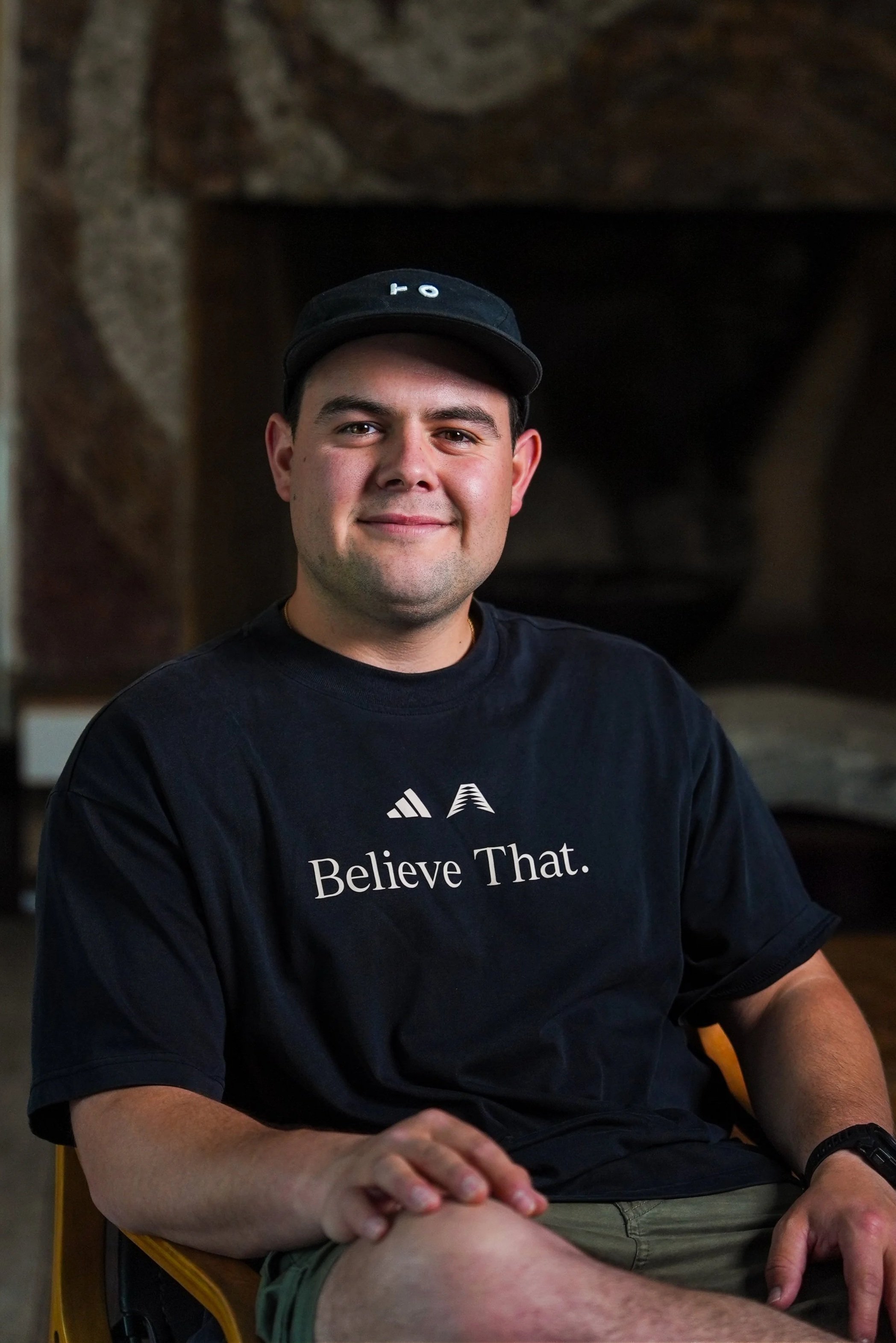 Joven hombre con gorra y camiseta negra que dice "Believe That", sentado en una silla de colores, en un ambiente interior con paredes de piedra.