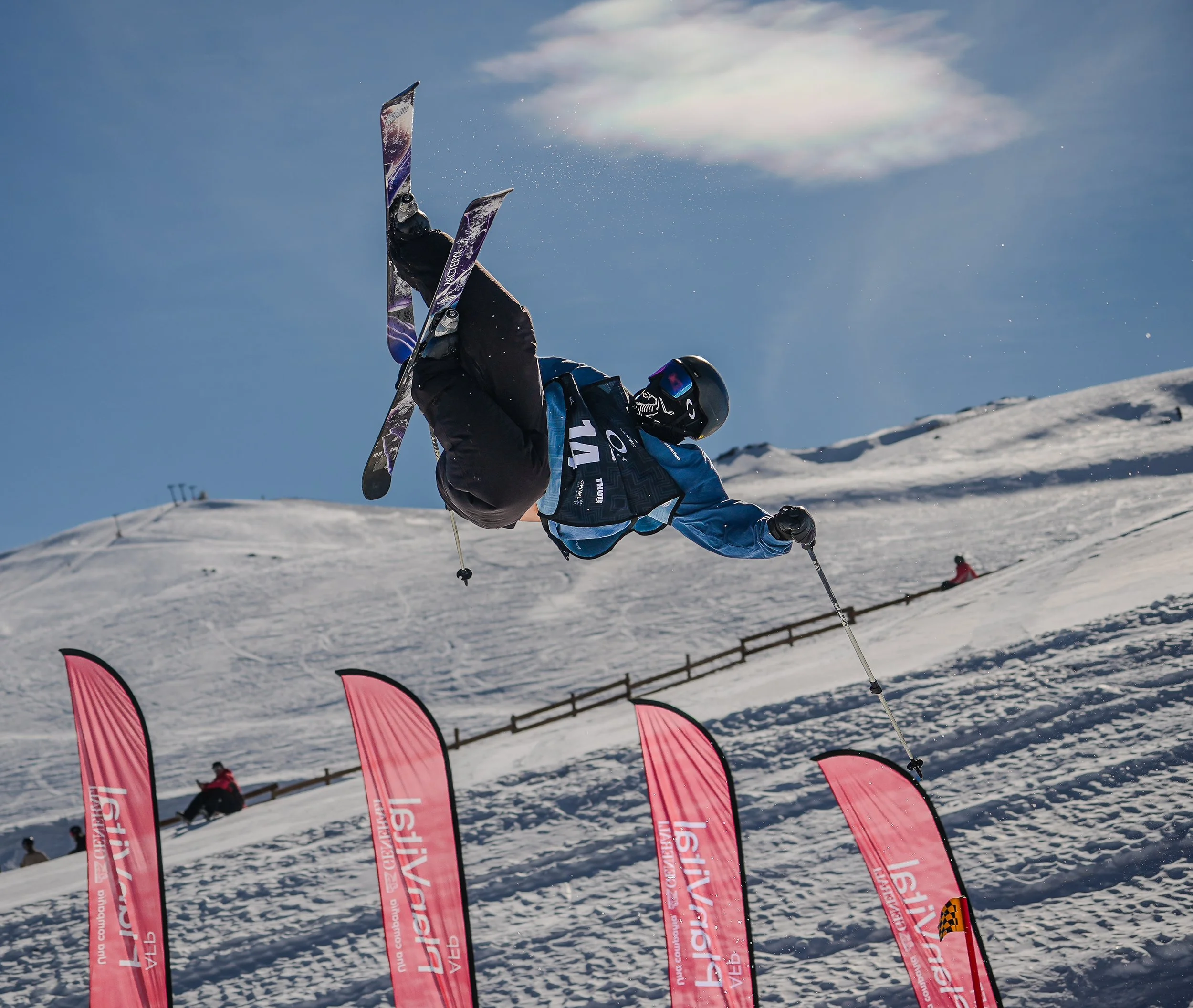Esquiador realizando una acrobacia en la nieve, con banderas rosas de fondo y cielo despejado.