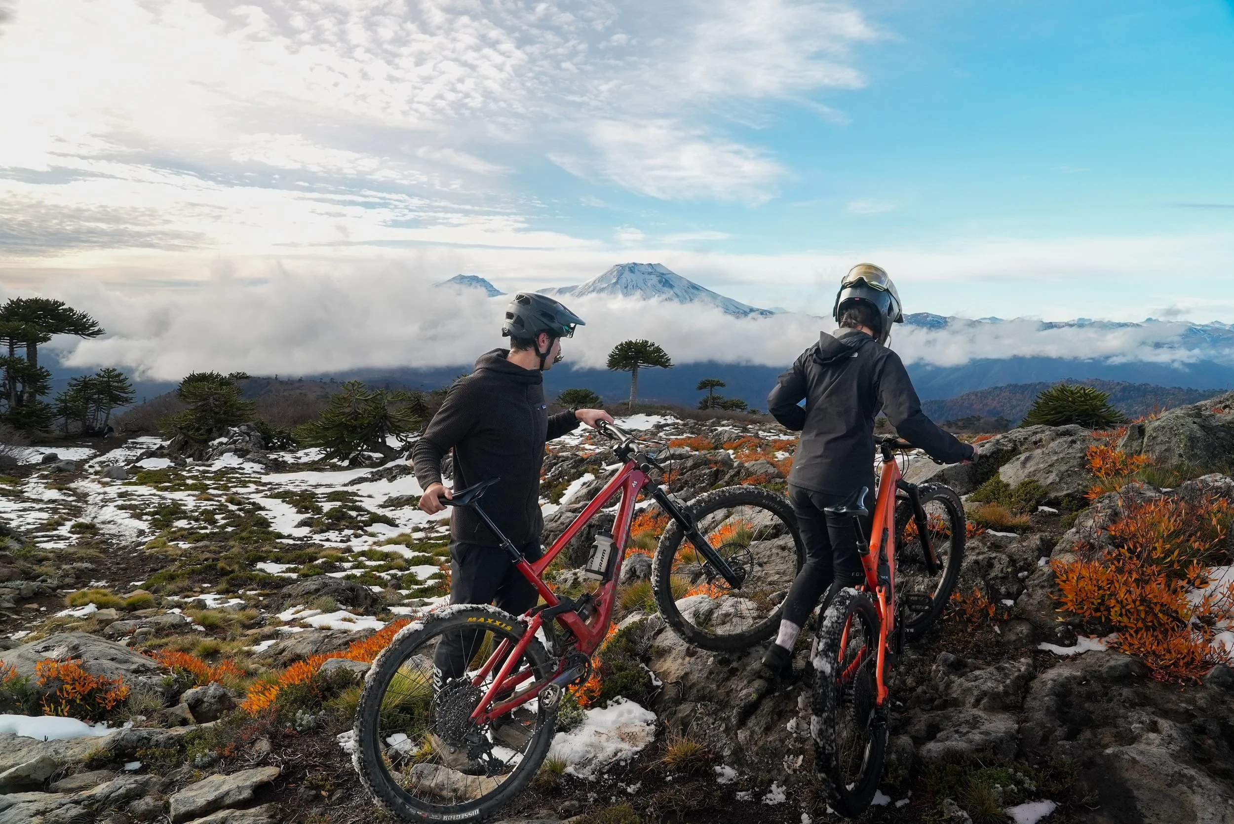 Dos personas con cascos de bicicleta en un paisaje montañoso con nieve, árboles y una montaña volcánica en el fondo, con nubes y cielo azul