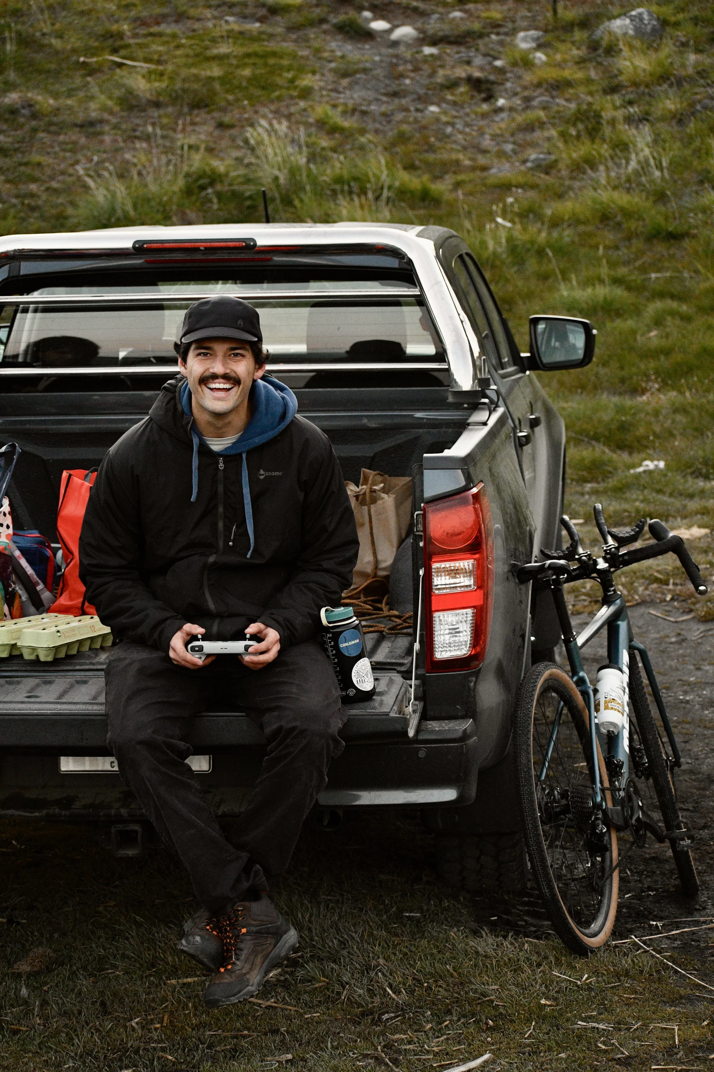 Hombre sonriendo sentado en la parte trasera de una camioneta, rodeado de bicicletas y equipo de camping en una zona natural con césped y arbustos.