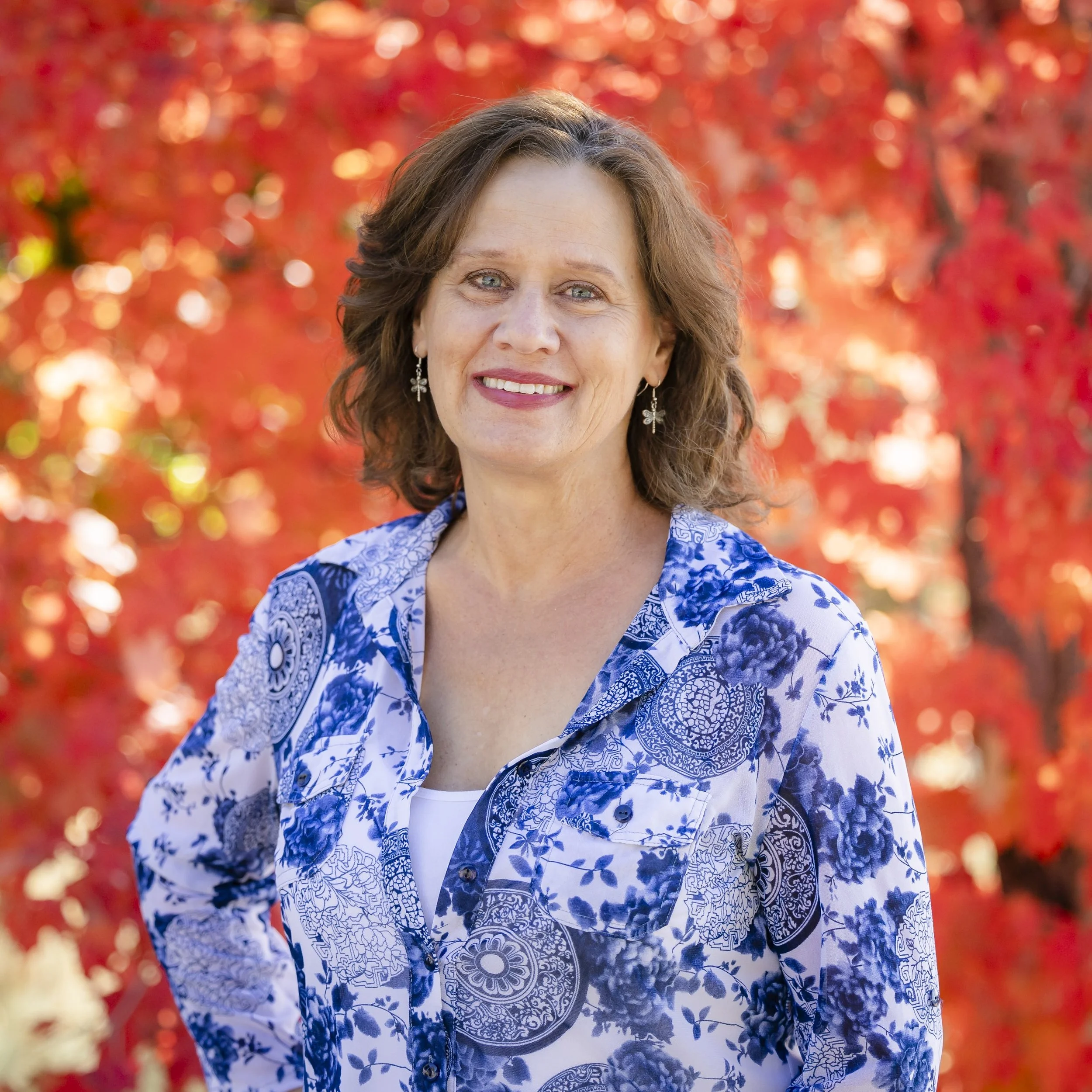 A woman with short, wavy brown hair and blue eyes, smiling, wearing a blue patterned top and earrings, against a dark gray background.
