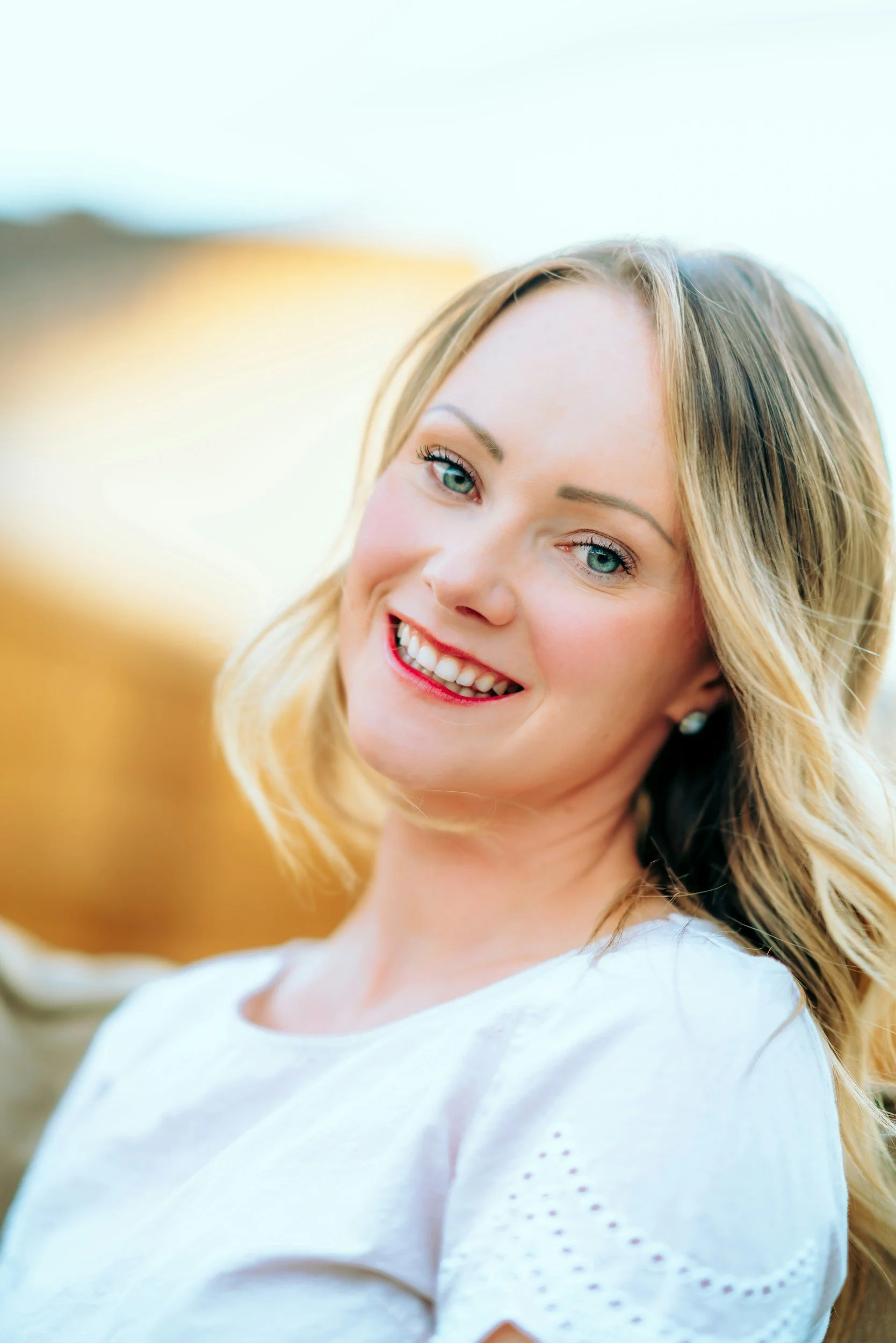 A smiling woman with blonde hair, blue eyes, and fair skin wearing a white top, standing outdoors on a sunny day.