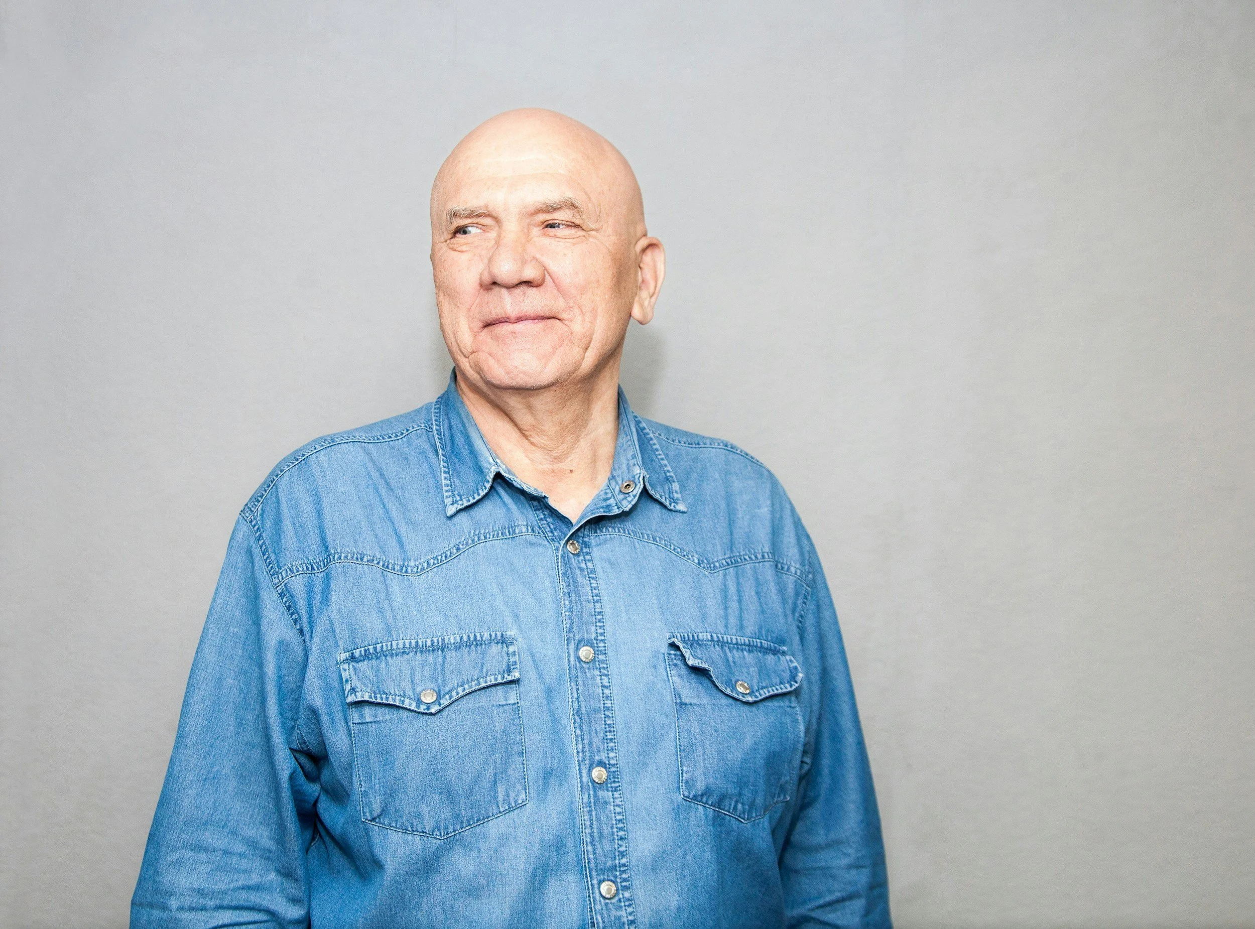 An older bald man wearing a denim shirt stands against a plain light gray wall, looking to his left with a slight smile.