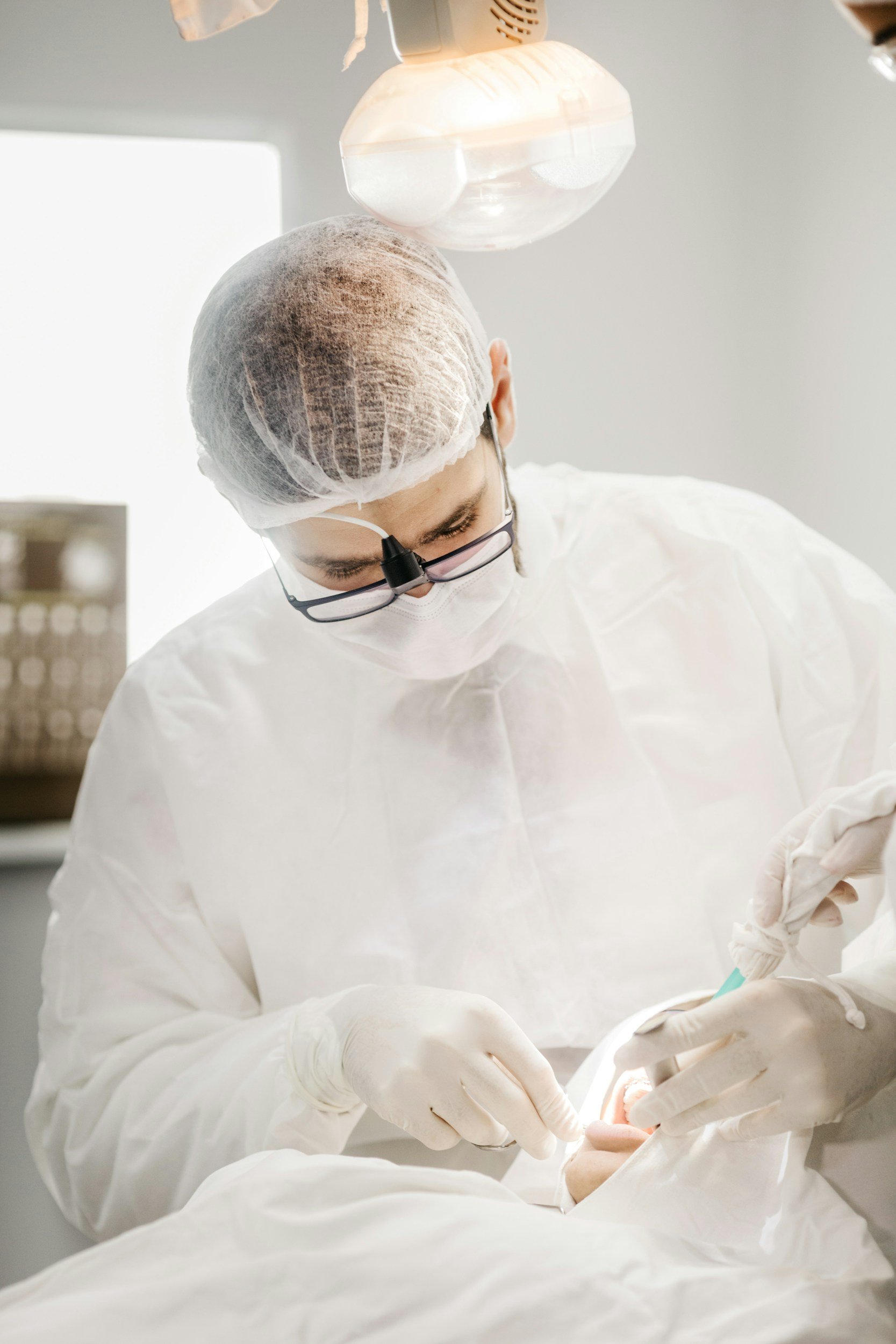 A medical professional wearing a hairnet, face mask, glasses, and gloves performing a procedure in a clinical setting.