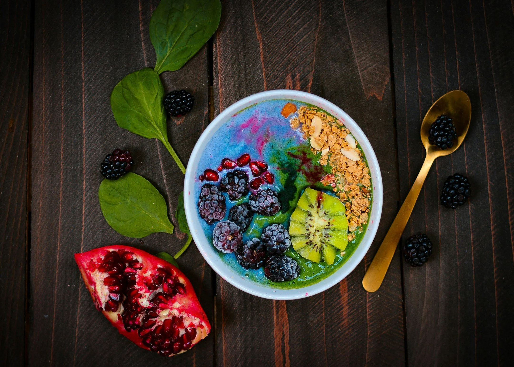 A smoothie bowl with colorful swirl topping, topped with blackberries, kiwi slices, granola, and almonds, on a dark wooden surface with scattered blackberries and a gold spoon with a blackberry.
