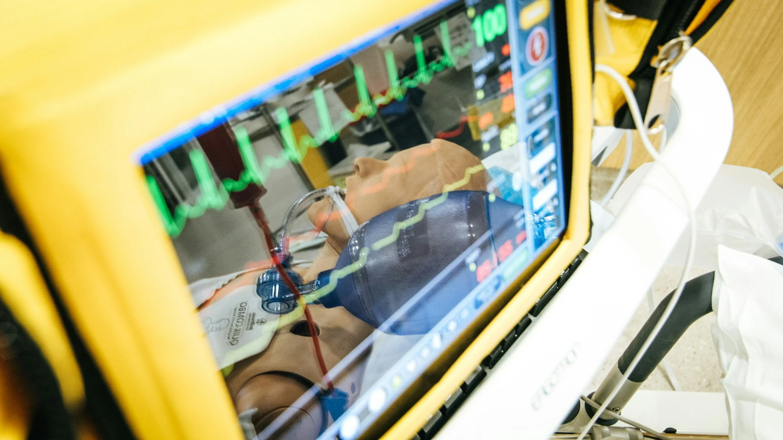 Person lying in a hospital bed connected to a monitor showing vital signs, viewed through the window of a yellow emergency medical device.