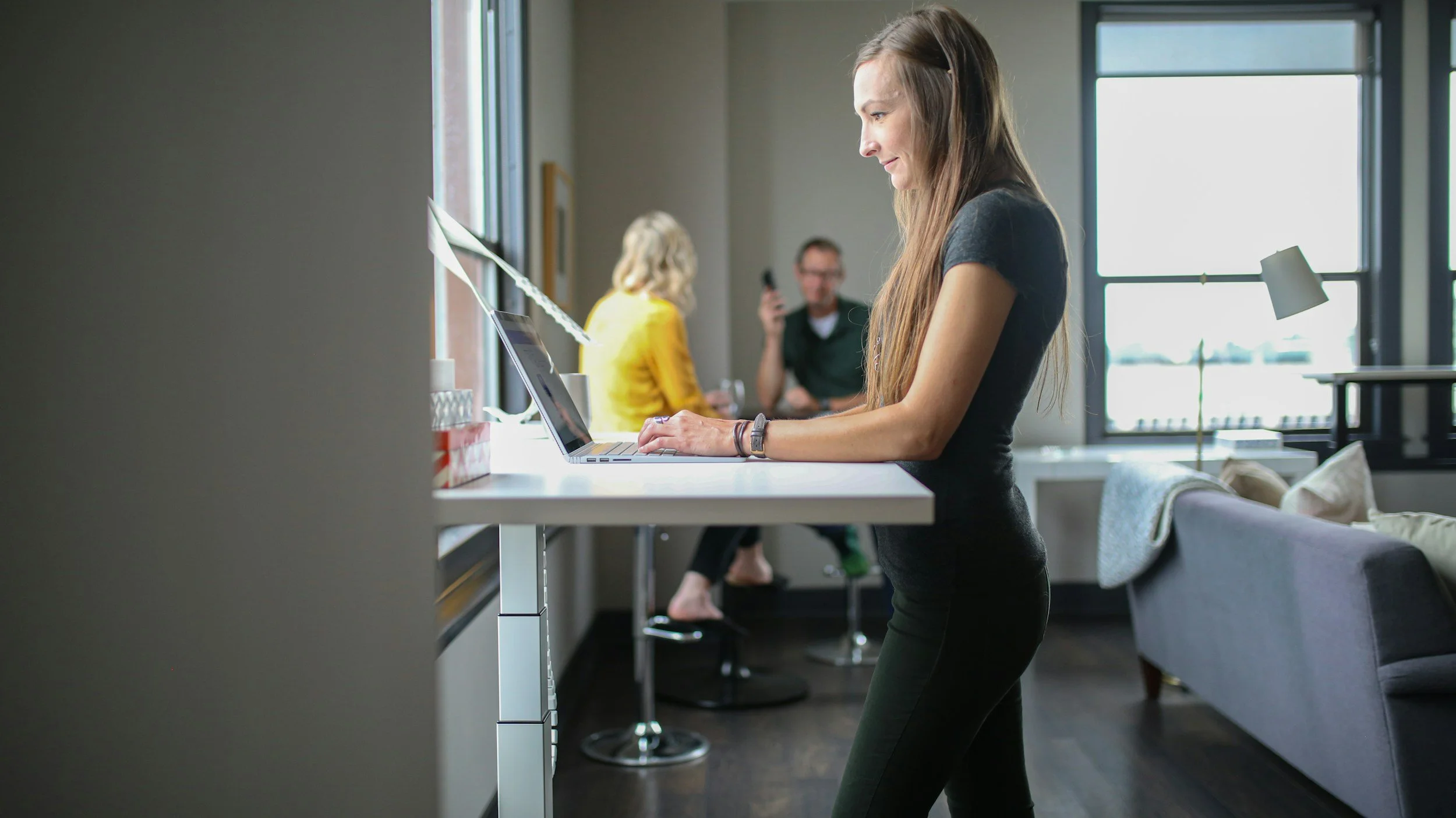 Woman standing at a high table working on a laptop in a modern, well-lit apartment with large windows. Other people are visible in the background socializing.