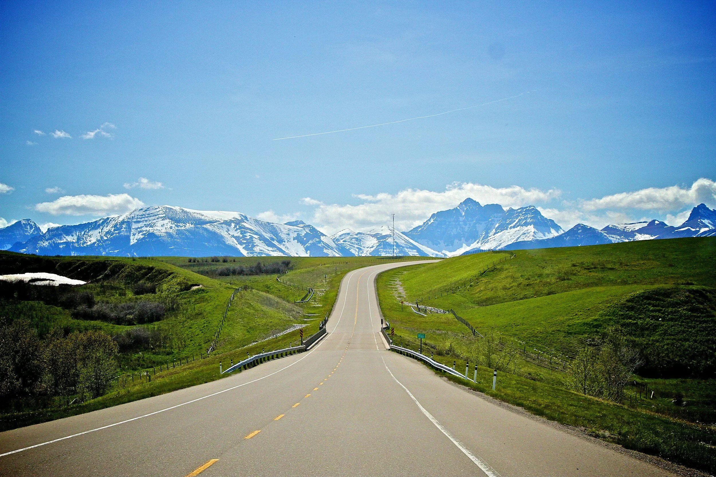 A winding road through green hills with snow-capped mountains in the distance under a blue sky.