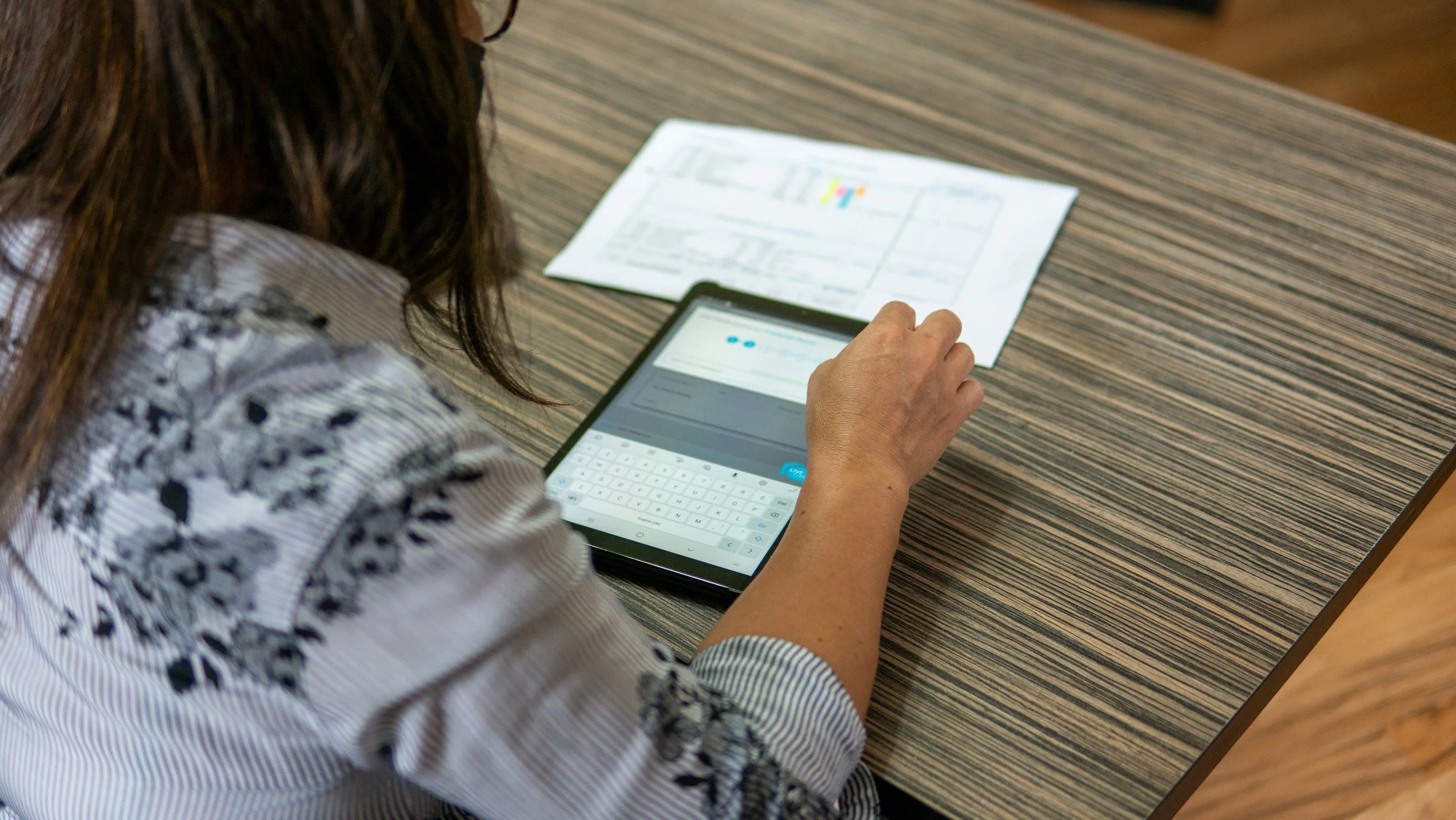 A person sitting at a striped table, using a tablet with a keyboard attachment, with printed documents on the table.