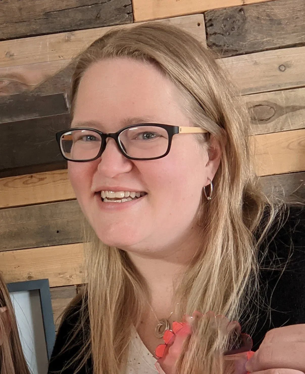 Close-up of a smiling woman with long blonde hair, wearing glasses and hoop earrings, holding a flower, with a wooden wall background.