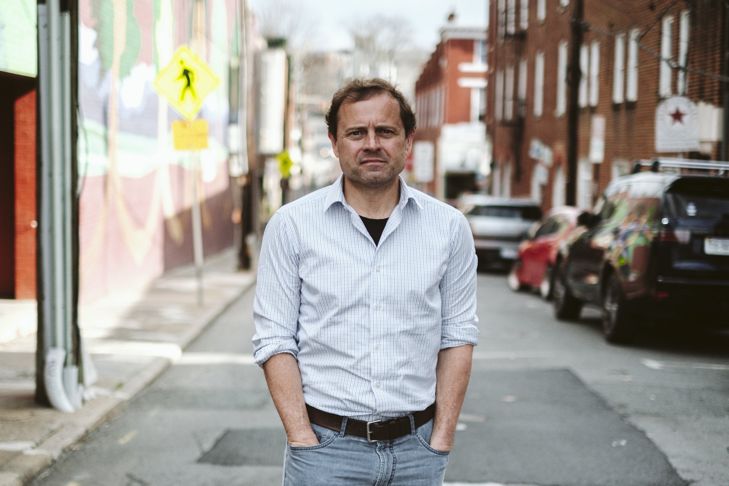 A man with brown hair, wearing a light blue dress shirt and jeans, walking on a city sidewalk with cars parked along the street and a painted mural on the building wall in the background.