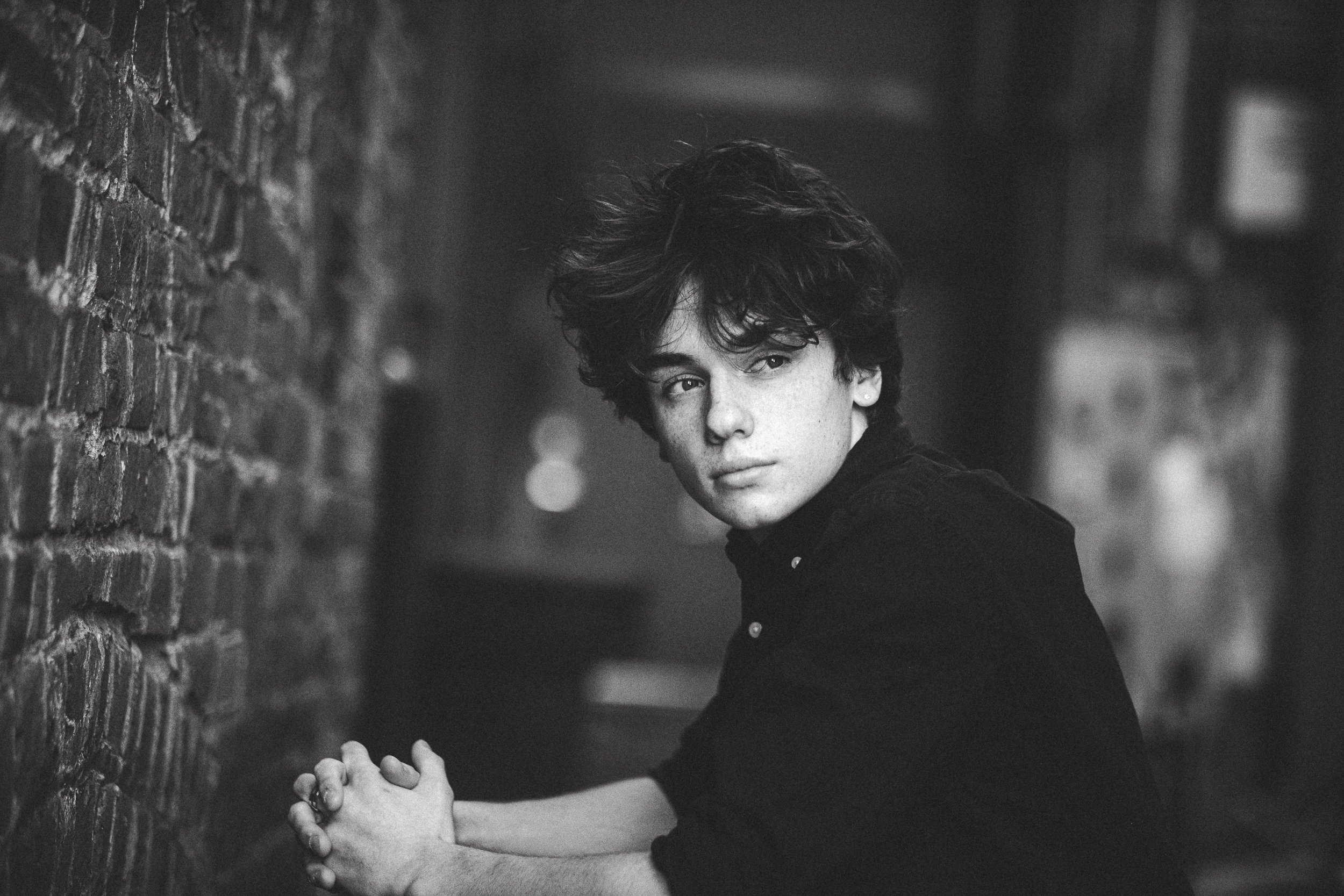 A black and white photo of a young person with short, curly hair, leaning against a brick wall and looking at the camera.
