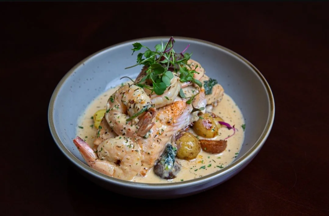 Seafood dish with shrimp and fish in a creamy sauce, garnished with microgreens, served with olives and vegetables in a gray bowl.