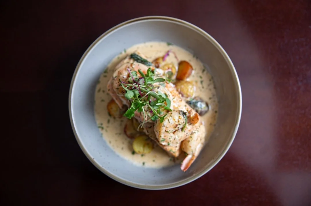 Bowl of creamy seafood dish with shrimp, garnished with herbs, served with vegetables in a white sauce on a dark wooden table.