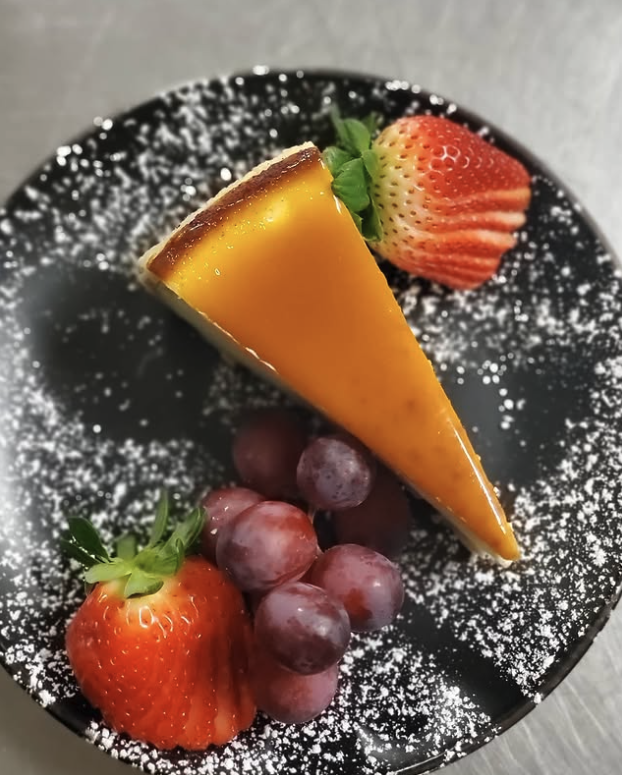 A slice of mango cheesecake garnished with strawberries, grapes, and a sprig of mint on a black plate dusted with powdered sugar.